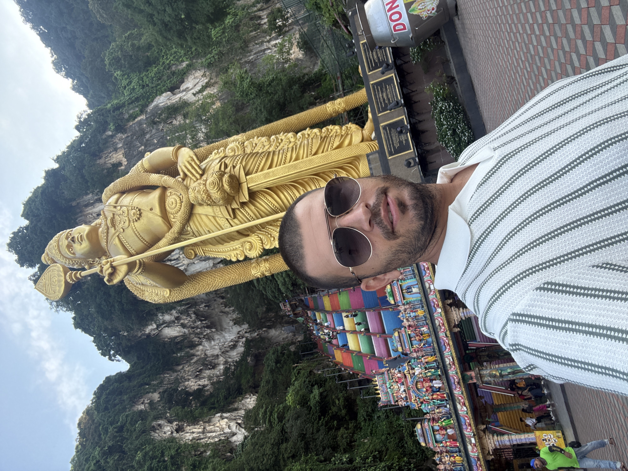 A man captures a selfie at a prominent religious and tourist site, showcasing a colossal golden statue and vibrant temple architecture. The man, positioned on the right side of the frame, wears sunglasses and a white polo shirt with thin dark stripes. To his left, a towering golden statue of a Hindu deity, likely Lord Murugan, stands prominently, holding a spear. In the background, steep, verdant hills covered in dense foliage rise under an overcast sky.
Below the golden statue and extending to the right, a multi-tiered, intricately decorated temple structure features numerous colorful steps, adorned with an array of smaller, brightly painted statues and carvings. Several other people can be seen near these colorful steps. In the upper right corner, a kiosk or stall is partially visible, displaying the text "DON" and some Asian script. The setting, with its distinctive golden statue, colorful temple steps, and limestone karsts, strongly indicates the Batu Caves in Selayang Municipal Council, Malaysia, during daytime.