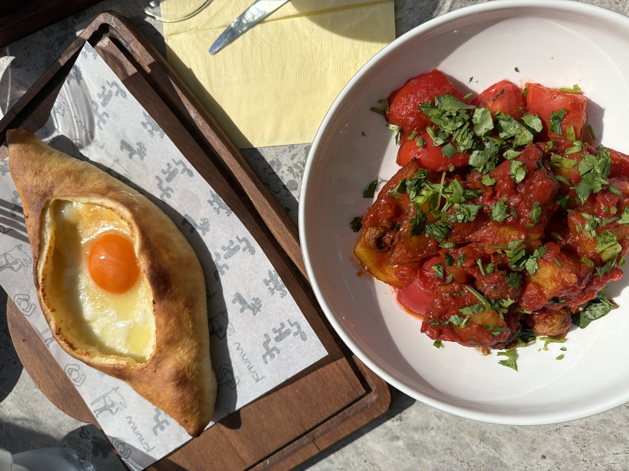 The image shows a table setting with two dishes. On the left, a khachapuri, a traditional Georgian bread filled with cheese and topped with an egg, is presented on a wooden tray lined with branded paper. The khachapuri has a characteristic boat shape with golden-brown crust. The filling consists of melted cheese and a partially cooked egg with a bright orange yolk.
On the right, a white bowl contains what appears to be a stew or a braised vegetable dish with a tomato-based sauce and fresh cilantro sprinkled on top. The dish seems to include tomatoes, and possibly potatoes or other root vegetables.
A yellow napkin and a knife are visible in the background. The setting suggests an outdoor dining experience, possibly in a restaurant or cafe.
