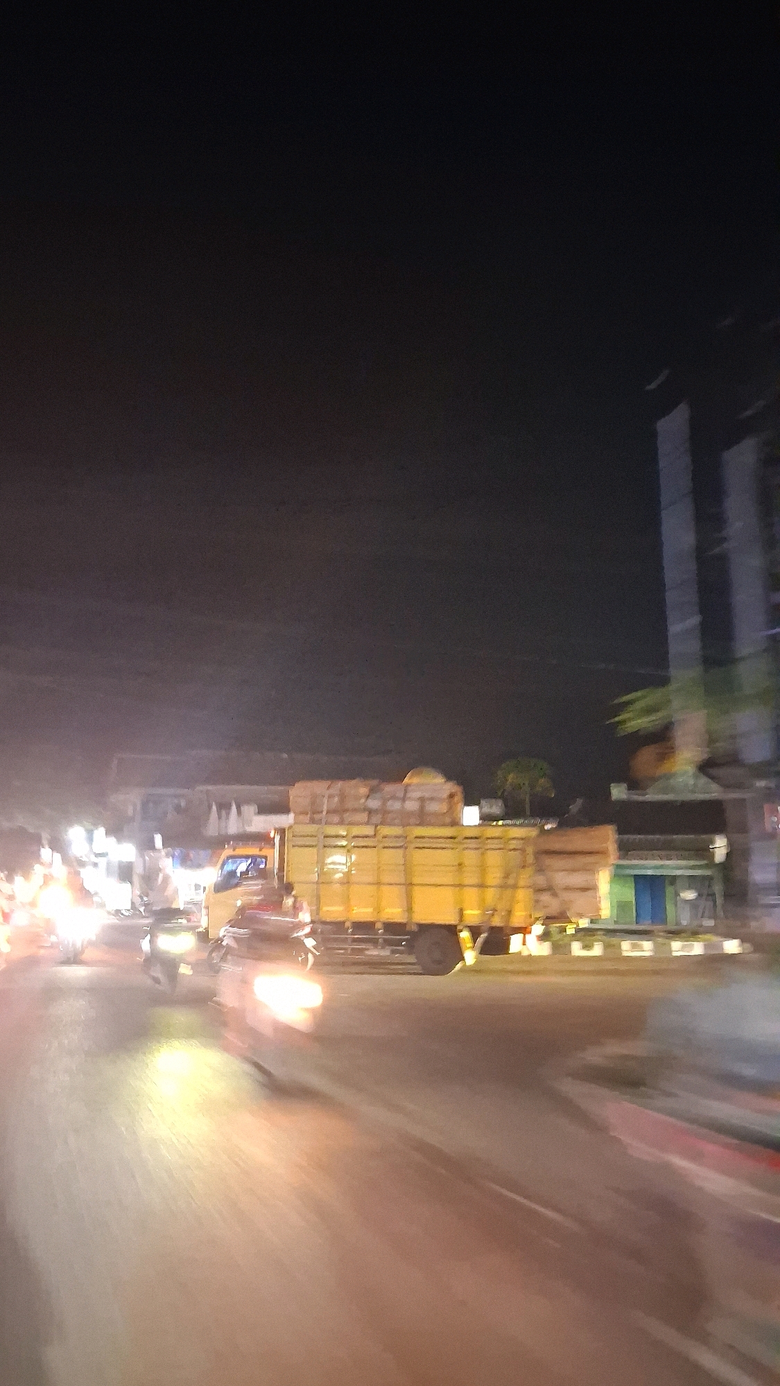 The image depicts a nighttime street scene in Berbah, Indonesia, characterized by motion blur and artificial lighting. A yellow truck carrying a load of what appears to be cardboard boxes is prominently featured in the middle ground, traveling on a road that curves to the right. The truck is illuminated by headlights from approaching vehicles and streetlights. Several motorcycles, their headlights also blurred due to motion, are visible on the road, indicating active traffic. In the background, building facades and a tall, dark structure, possibly a high-rise, are partially discernible. The overall impression is one of a busy urban environment at night, with vehicles in motion on a paved road.