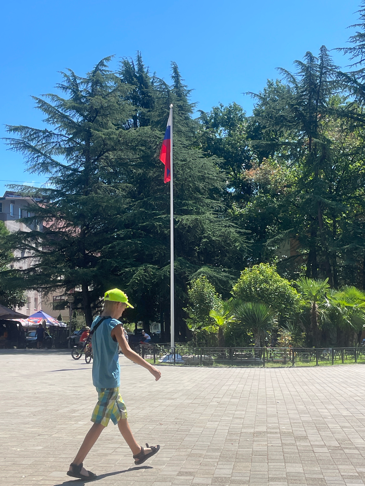 A young boy wearing a bright yellow baseball cap, a blue tank top, and checkered shorts walks across a paved plaza on a sunny day. He is in the foreground, facing to his right, with his left leg extended forward and his right leg behind him, suggesting he is mid-stride. He wears dark sandals. In the background, a Russian flag flies from a tall flagpole against a clear blue sky. Large evergreen trees and other lush vegetation are visible behind the flag and on the right side of the image. To the left of the flag, a building with a reddish-brown roof can be seen. Further left, a colorful tent structure and some people are present, along with a bicycle. The plaza is paved with rectangular gray tiles. The scene appears to be an outdoor public space in Sochi, Russia, likely a park or square with significant greenery and a backdrop of urban architecture.