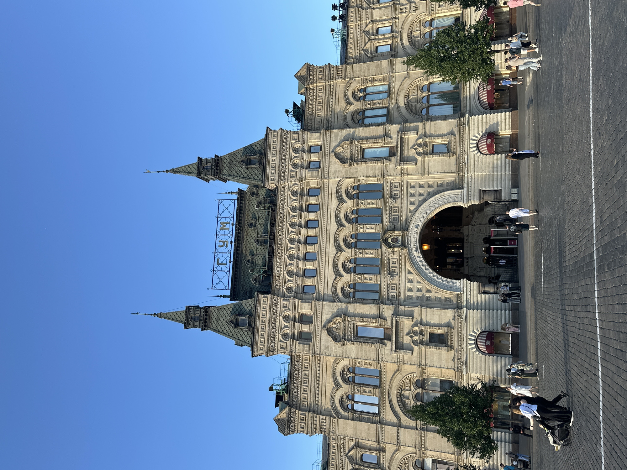The image shows the GUM department store, a prominent landmark located on Red Square in Moscow, Russia. The building is a large, ornate structure with a light-colored facade, featuring multiple arched windows and decorative stonework. Two prominent towers with green roofs rise from the building's main structure, topped with spires. The letters "ГУМ" (GUM) are visible on a sign above the central section of the building. The sky above is clear and blue. In front of the building, a cobblestone plaza is visible, with several people walking around. Some are strolling, while others are pushing strollers or talking in groups. There are also trees and awnings in front of the store entrances. The scene appears to be an outdoor public space with light foot traffic during daylight hours.