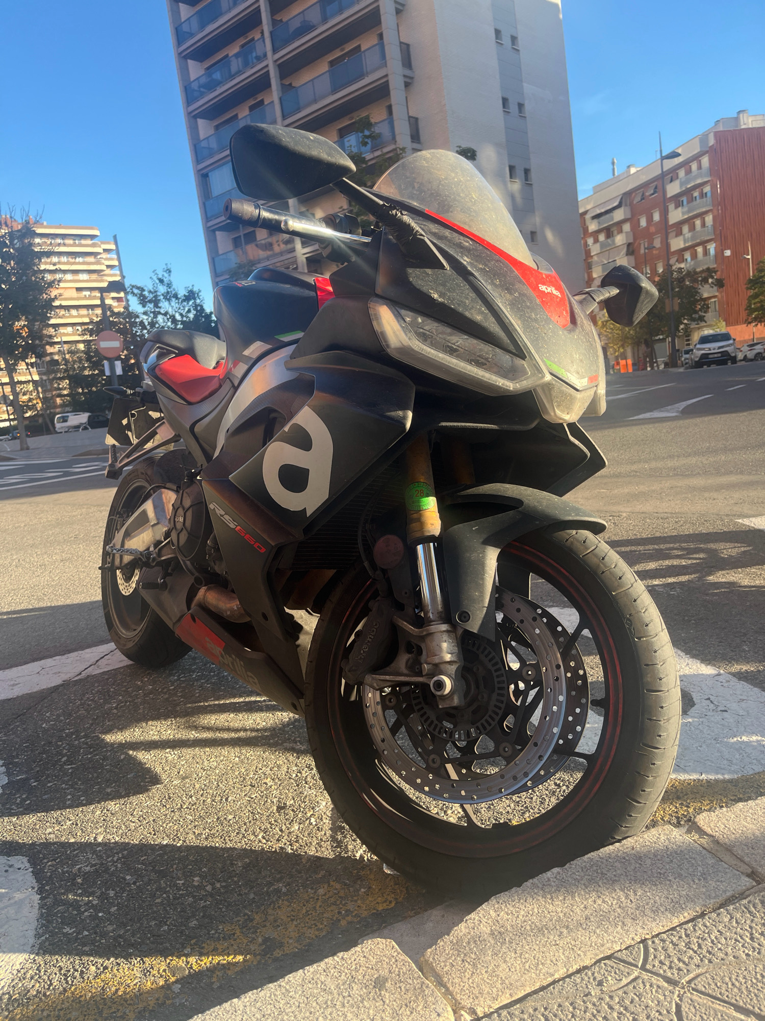 A black and red Aprilia RS 660 motorcycle is parked on a paved street, possibly in Zona Eixample, Spain, under a clear blue sky. The motorcycle is positioned at an angle, facing towards the viewer's right, with its front wheel on a crosswalk marked with white and yellow lines. The motorcycle is detailed with an "a" logo on its side, the "Aprilia" brand name on the fairing, and "RS660" lettering on the lower fairing. The bike appears dusty, particularly on the front fairing and windshield. In the background, several multi-story apartment buildings with balconies and windows are visible. Trees line the street, and there are traffic lights and street lamps. The overall scene suggests an urban setting during daylight hours.