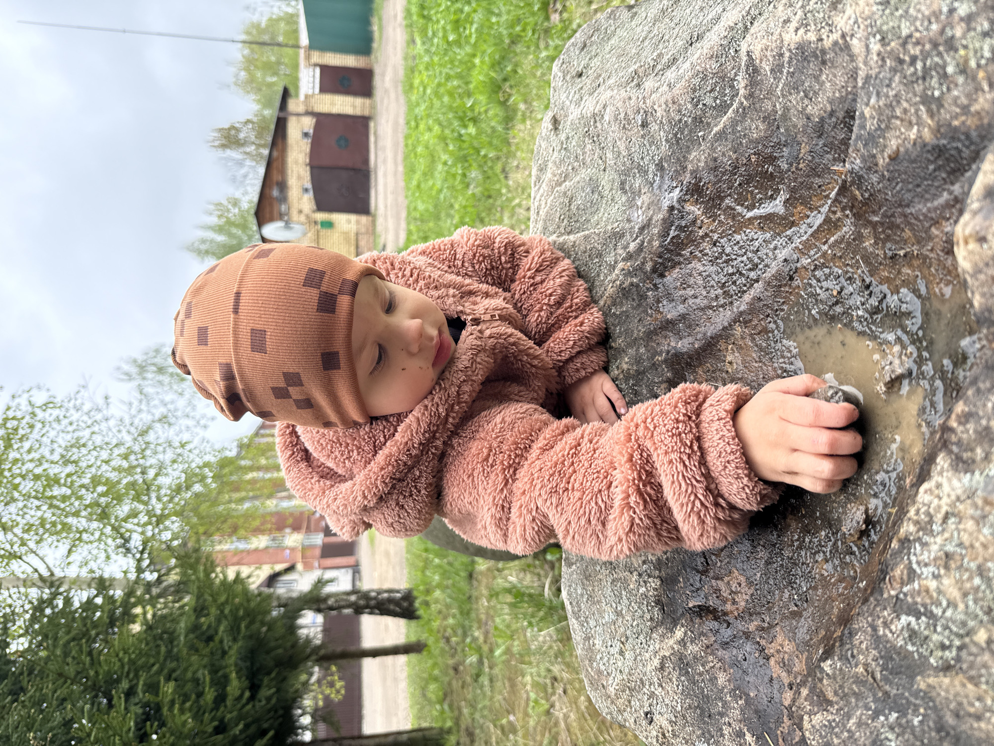 A young child wearing a brown, checkered hat and a fuzzy, brown jacket is lying on a large, gray rock in a grassy area. The child's right hand is immersed in a puddle of muddy water on the surface of the rock. In the background, there are buildings, including a garage with multiple roll-up doors, and green trees. The scene appears to be outdoors, possibly in a park or backyard, but the specific location in the city of Unknown, Unknown is not discernible. The photograph is rotated to the left, which affects the initial perception of the scene's orientation.