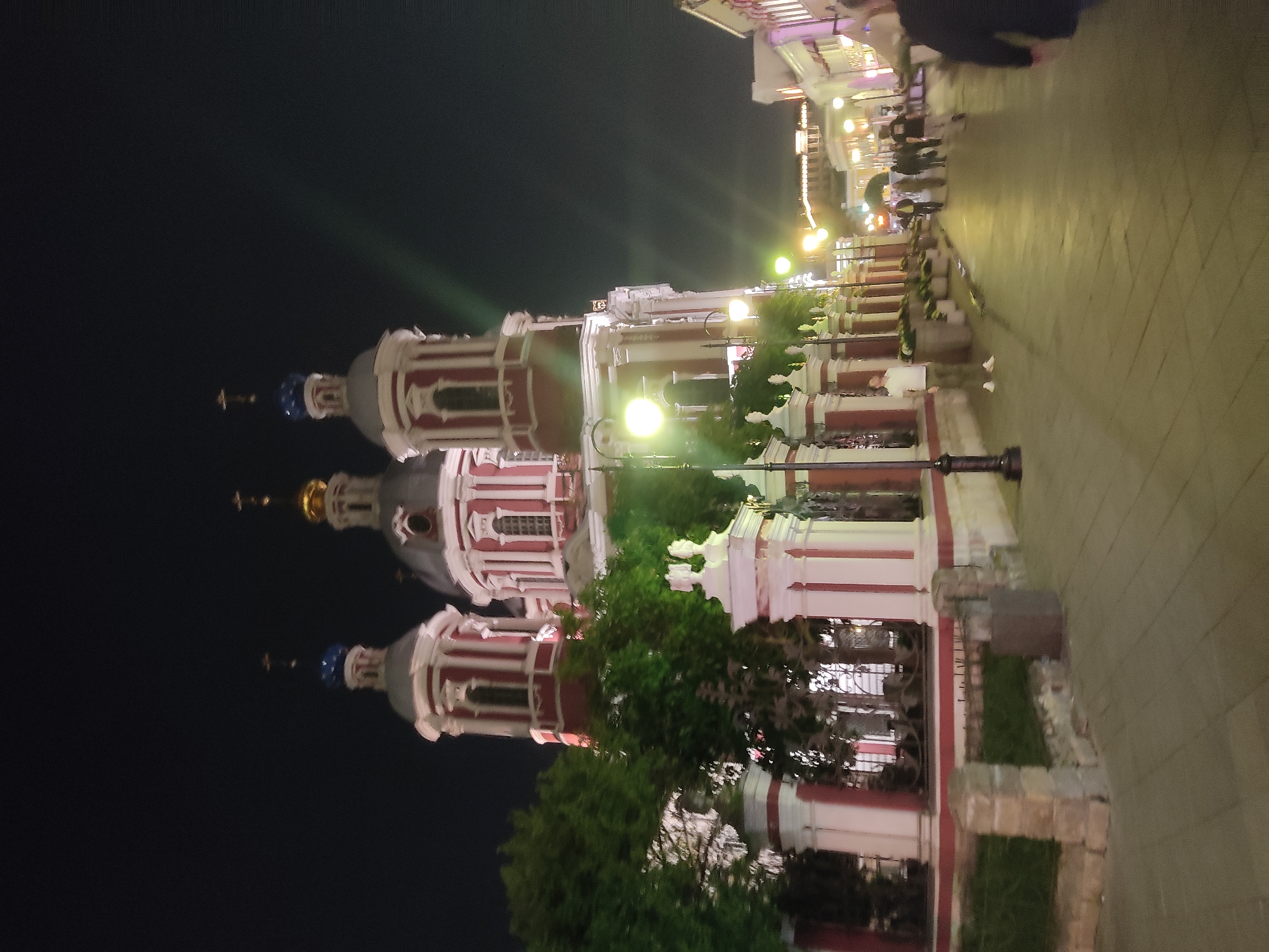 Here is a detailed description of the image:
The image shows the Cathedral of the Icon of the Mother of God "Joy of All Who Sorrow" on Bolshaya Ordynka Street in Moscow, Russia, at night. The cathedral features three domes, each topped with a cross. The domes are painted in a red and white striped pattern, with the lowest dome bearing a blue roof and a blue spire, the middle dome a dark grey roof and a golden spire, and the topmost dome a dark grey roof and a blue spire.
Along the right side of the frame is a paved walkway illuminated by ornate street lamps. A wrought iron fence separates the walkway from the cathedral grounds, which are adorned with shrubs and trees. The walkway has multiple people walking along it. The sky is completely black, emphasizing the illuminated cathedral and the street scene.