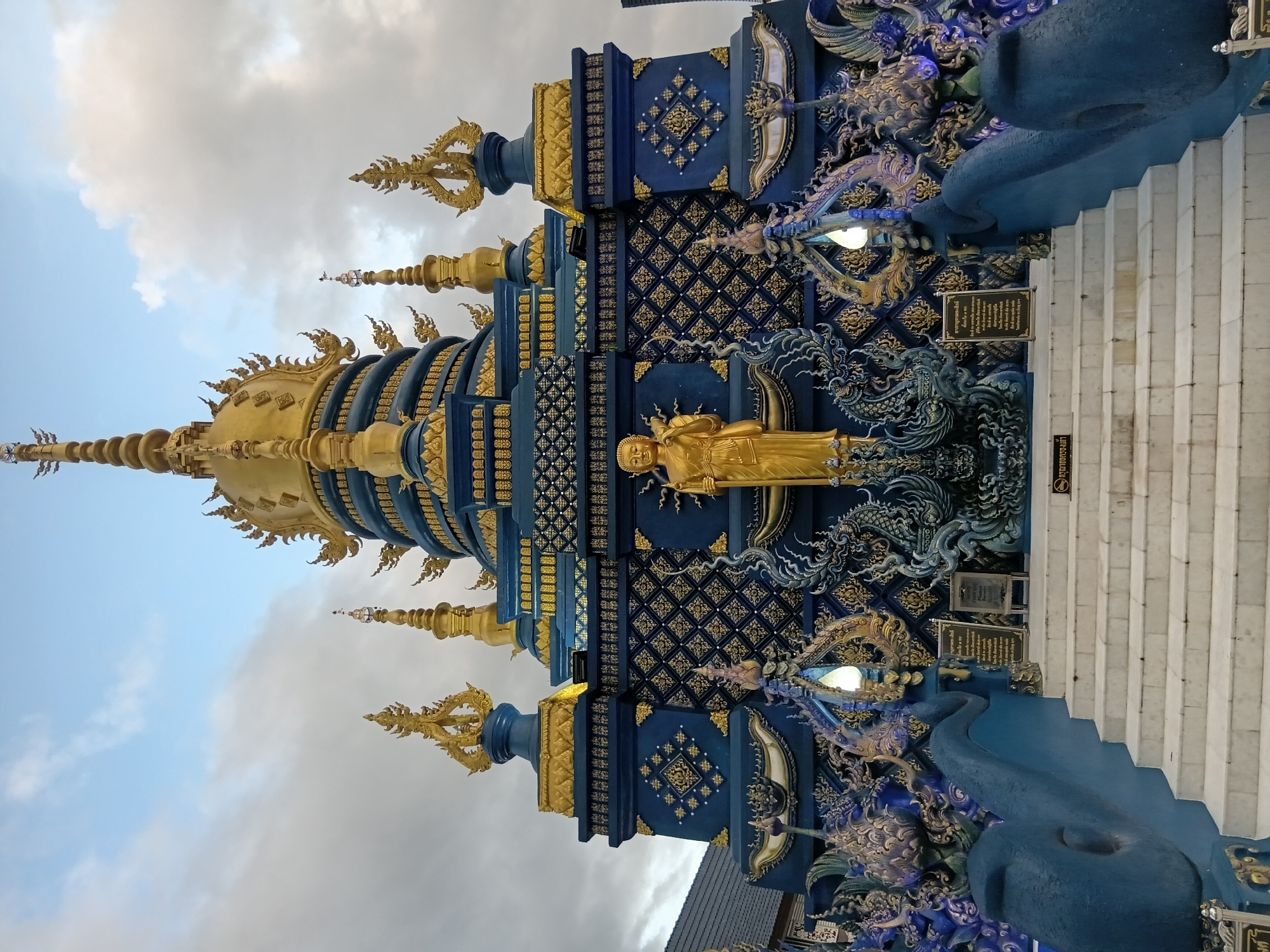 The image displays the Wat Rong Suea Ten, also known as the Blue Temple, located in Chiang Rai City Municipality, Thailand. The temple is characterized by its vibrant blue and gold architecture, featuring intricate decorative patterns and ornate spires. A golden statue of Buddha stands prominently in front of the temple's facade, which is adorned with mythological creatures and celestial motifs. White marble steps lead up to the temple entrance. The sky above is partly cloudy.