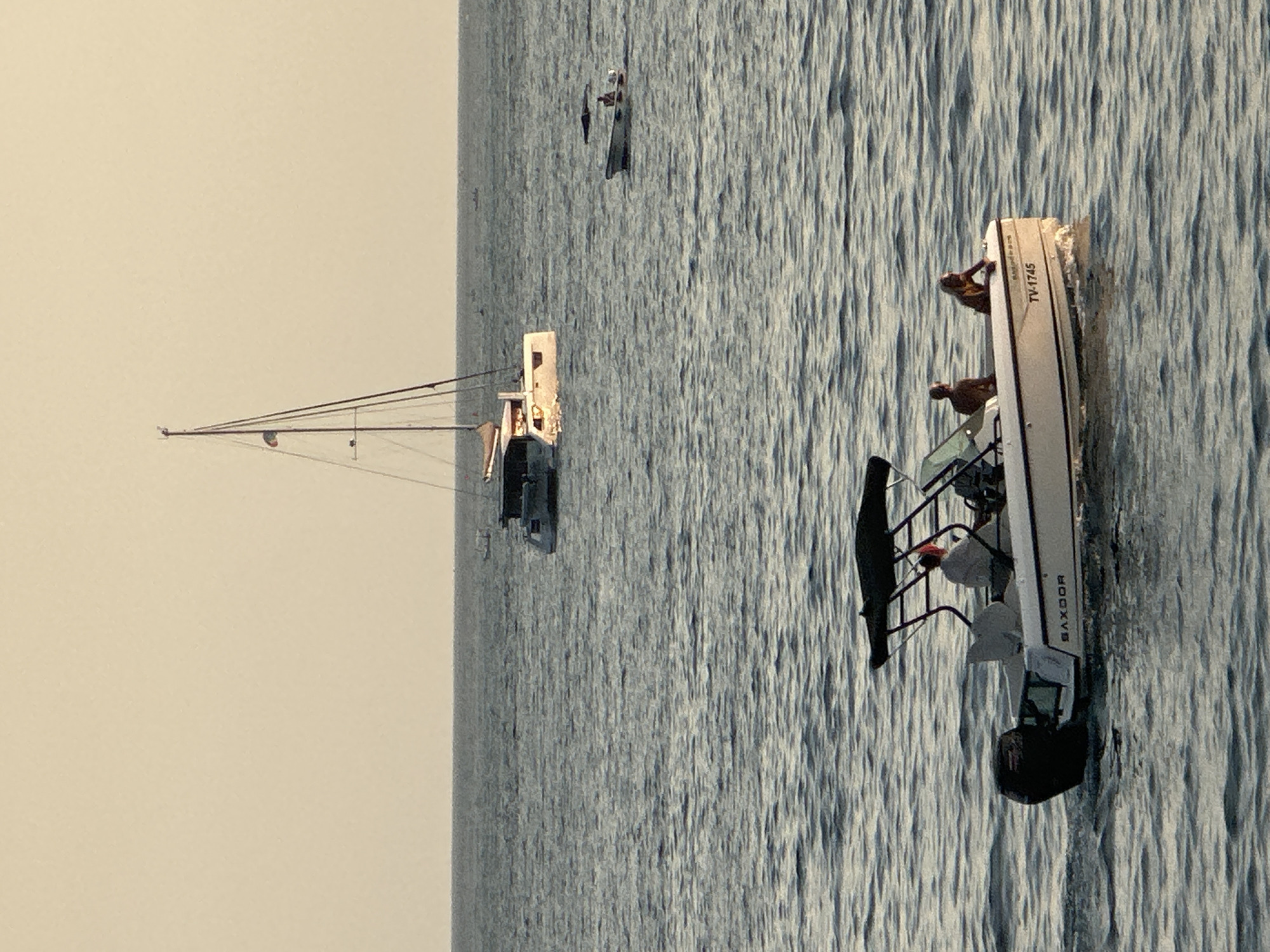 A wide-angle, eye-level shot captures a serene marine scene with multiple vessels on the water. The predominant color palette is dominated by the soft, muted tones of the sea and sky, hinting at either dawn or dusk. In the foreground, a white speedboat is cruising, with three individuals on board. The boat features a black canopy and a visible outboard motor. A name or logo, "SAXDOOR," is present on the side of the boat, as well as a license plate reading "TV-1745." Further in the distance, a white catamaran with two hulls and a tall mast is anchored. This vessel appears to be a sailboat, with a flag visible on the mast. Another smaller, white boat is also visible in the middle ground, with a single person onboard. The water is calm with gentle ripples, and the horizon line is clearly defined. The scene is set in the waters off the coast of Budva, Montenegro.
