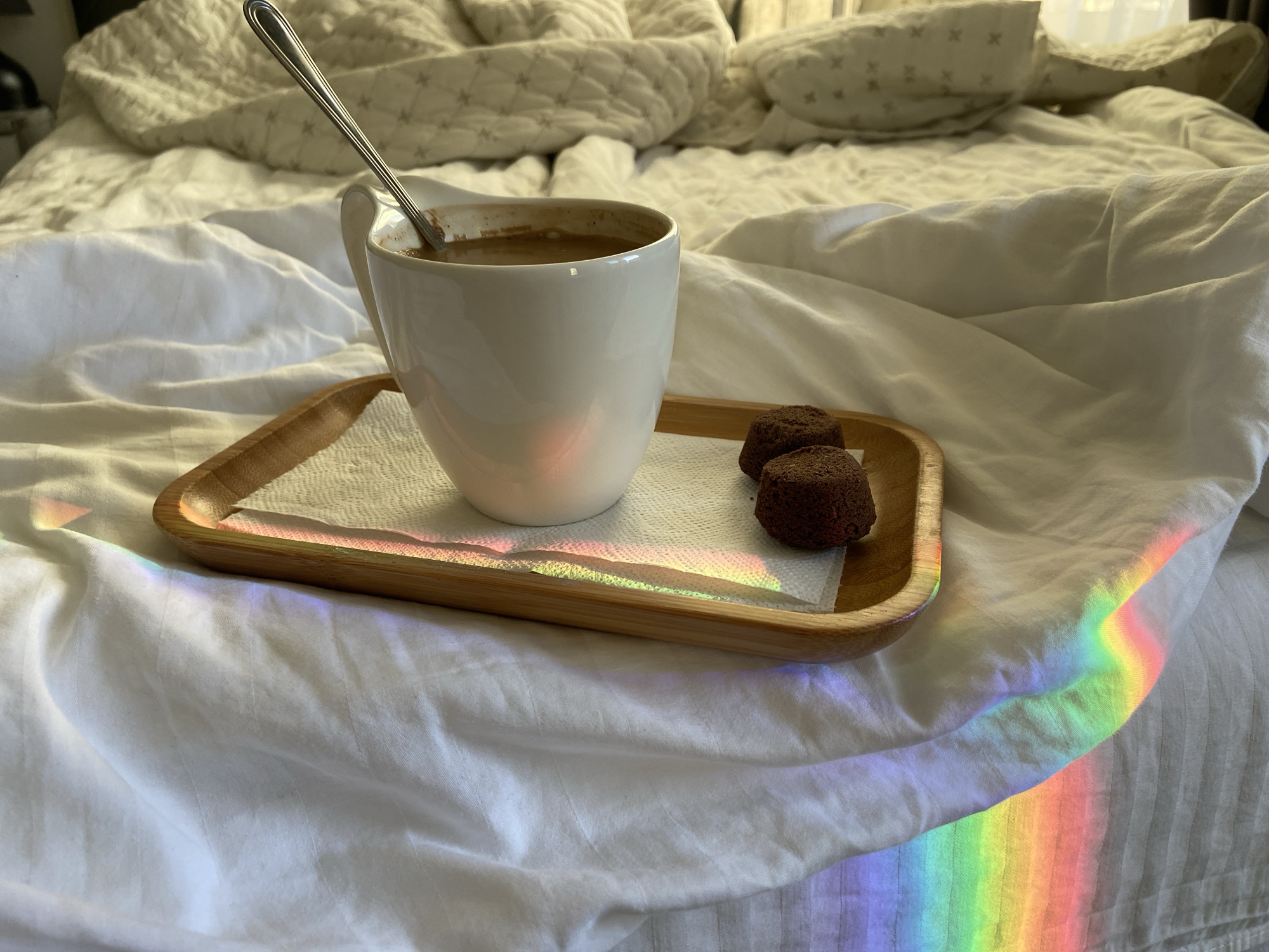 A close-up shot depicts a wooden tray resting on white linens, likely in a bedroom. The tray holds a white mug containing coffee with a spoon inside, along with two small, round, dark brown treats, possibly cookies or brownies. A rainbow refraction casts light across the white linens. The location is not discernable from the image and the city of Budva, Montenegro cannot be determined.
