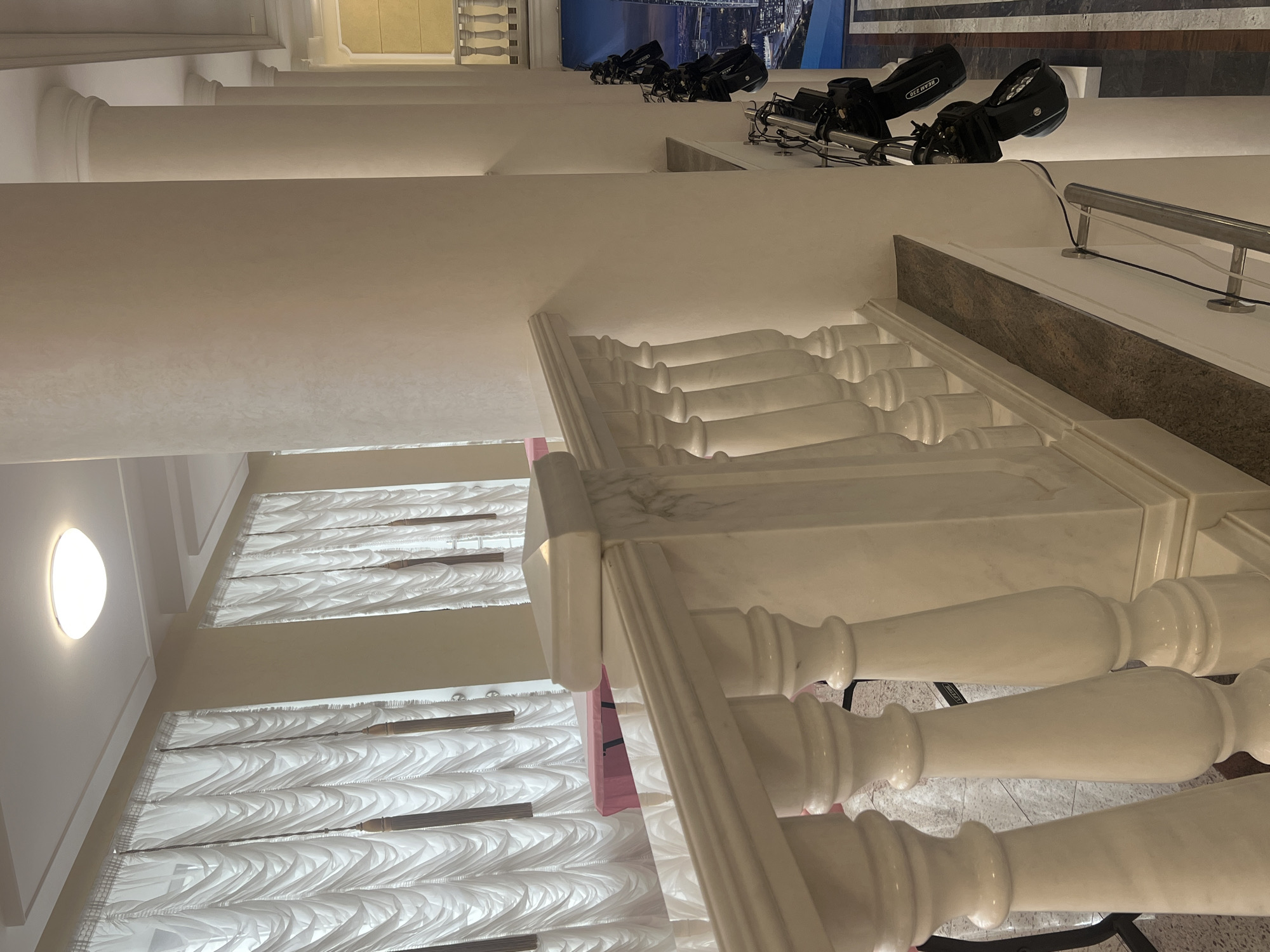 This image captures the interior of a large, elegant building, likely a hotel lobby, event hall, or theater foyer, located in Votkinsk, Russia. The central focus is a grand staircase featuring white marble balustrades with ornate, turned spindles and a dark, polished marble capping on its railing.
To the left, a series of tall, classical white pillars or columns stand prominently, creating an imposing architectural line. The walls are a uniform light beige or off-white, with some sections displaying a subtle, textured finish. The space is illuminated by various artificial light sources, including recessed strip lighting along ledges and architectural details, and a distinct, round ceiling light fixture with a warm glow in the lower left corner.
On an upper level, several black theatrical lighting fixtures, resembling moving head lights or stage lights, are positioned. Cables are visible running from these units. One of these fixtures clearly displays the text "BEAM 230." These lights appear to be aimed towards a large, dark-toned graphic or screen on an opposing wall, which depicts a nocturnal city skyline. A simple, silver metal handrail is also present on this upper level.
Below the upper level, multiple windows are covered by white, intricately pleated or textured curtains/blinds, adding a decorative element. A thin, flat pink object is partially visible near the base of the staircase railing, possibly a sign or part of a temporary setup. The absence of people suggests the space is currently unoccupied or being prepared for an event. The overall atmosphere is one of formal grandeur combined with modern event-hosting capabilities.