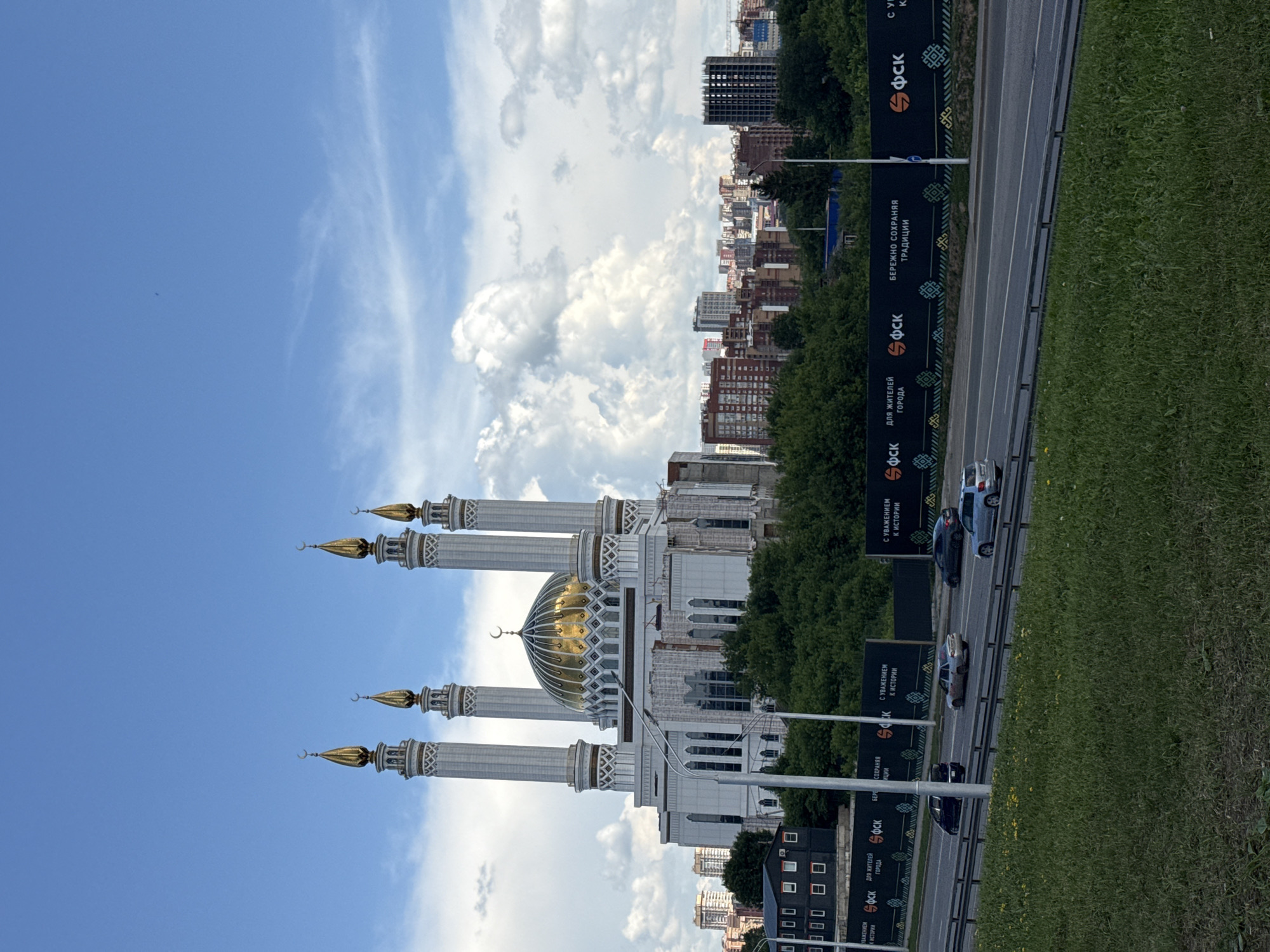 A wide-angle, eye-level, outdoor, daytime shot captures the side of a grand mosque with a golden dome and four tall, white minarets topped with golden crescents, set against a bright blue sky with scattered white clouds. The mosque appears to be under construction, with scaffolding visible on its facade. Below the mosque, a dense line of green trees forms a natural barrier. Beyond the trees, a cityscape of multistory buildings, including apartments and a dark, unfinished skyscraper, stretches across the horizon. In the foreground, a multi-lane road runs parallel to a grassy embankment. Several cars are driving on the road, and a dark fence with white text and decorative patterns runs along the side of the road.