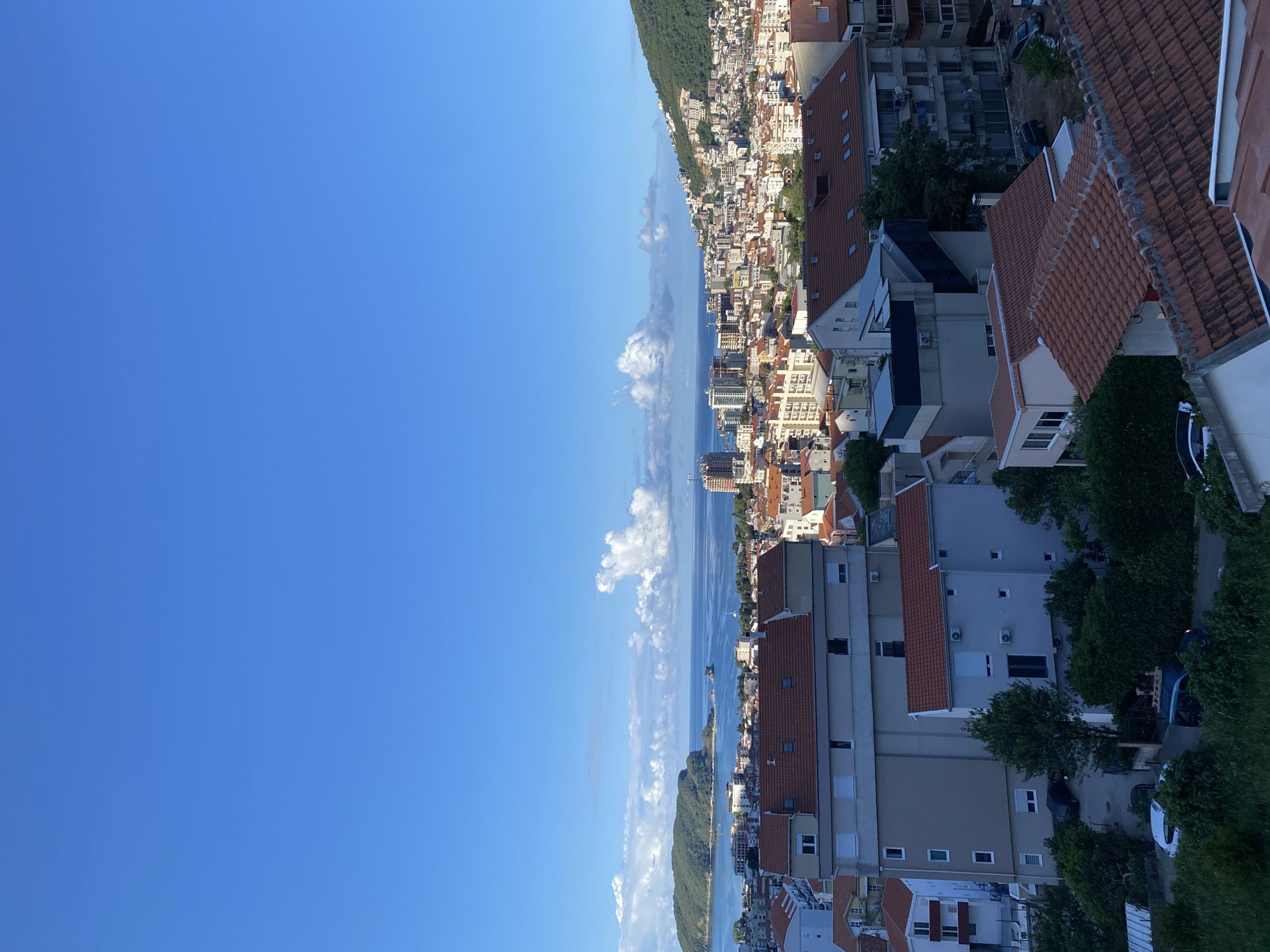 The image shows a view of Budva, Montenegro, from an elevated position. A clear, blue sky occupies the left half of the image. The Adriatic Sea, speckled with white clouds reflecting on its surface, runs vertically down the center of the image. Along the coastline, a dense collection of buildings with orange-tiled roofs and light-colored walls make up the cityscape. A forested mountain range is visible along the upper portion of the coastline. In the right half of the image are closer buildings, likely residences, with similar architectural features and glimpses of trees and parked vehicles. The image appears to be taken during daylight hours with ample sunshine.