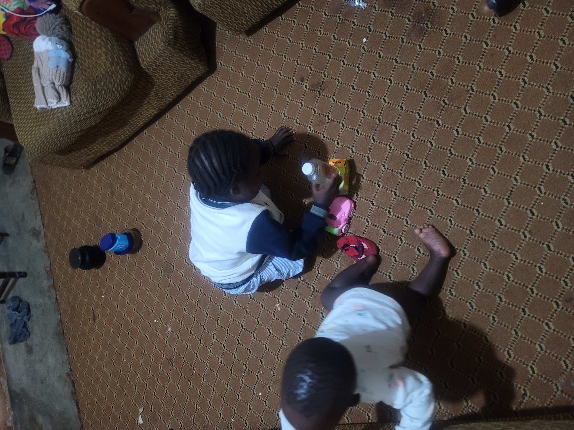 Two young children are on a brown patterned carpet, one standing and one sitting. The standing child, facing away, is wearing a white long-sleeved shirt and dark pants, with one red sandal on their left foot. The sitting child, facing towards the right and slightly downwards, is wearing a white long-sleeved shirt with blue trim and dark blue pants. The sitting child is reaching for a bottle with a white cap and a yellow and orange box, and there are two pink shoes nearby. To the right of the children, there are two blue and black containers and a dark grey sock on the floor. A brown sofa with a textured fabric is partially visible on the right side of the image, with a knitted glove on its armrest. The lighting suggests an indoor environment, possibly a living room, with a shadowed area indicating a light source from above or to the side.