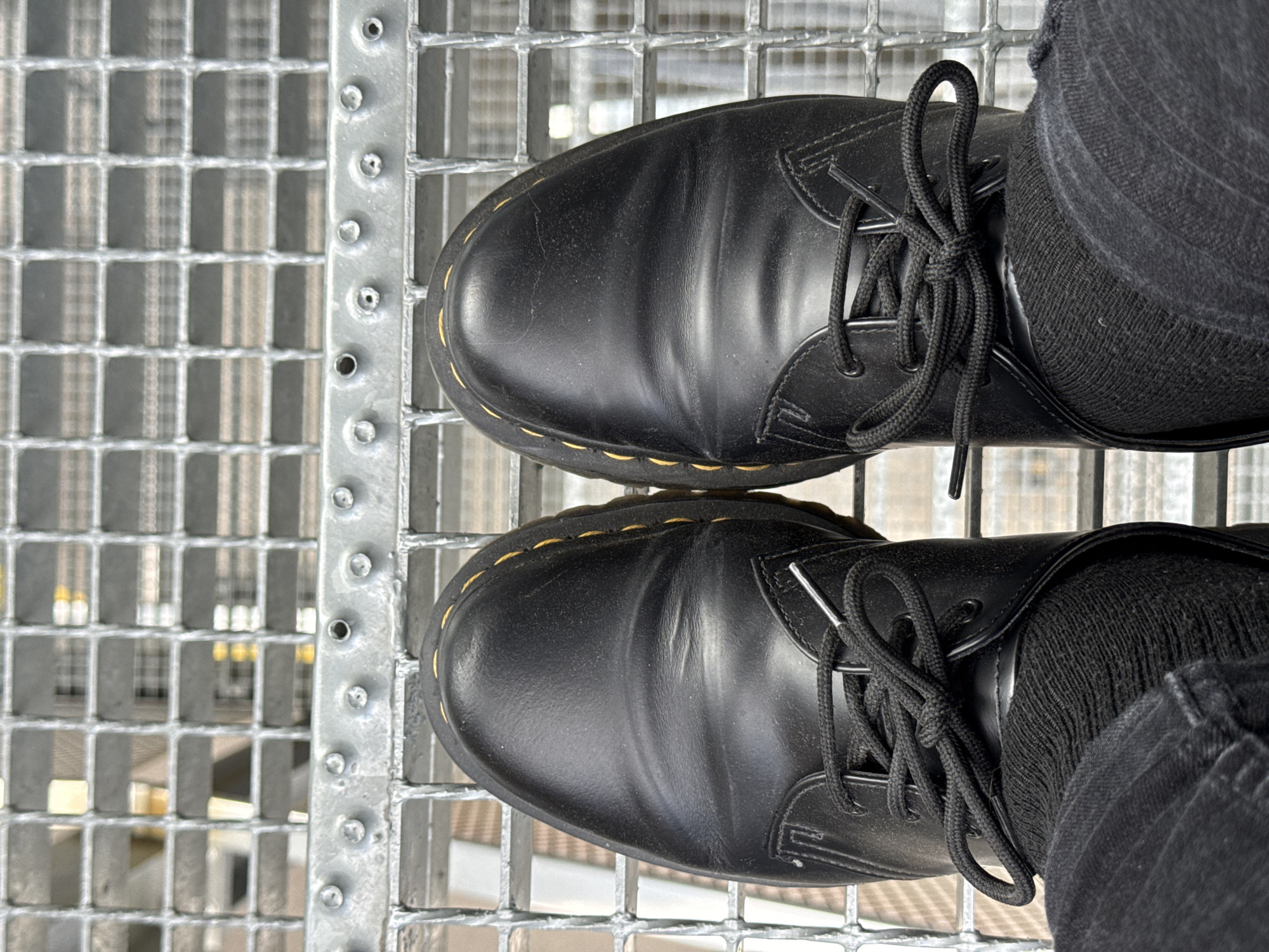 This is a close-up, top-down view of two black leather lace-up boots, likely Dr. Martens, resting on a metal grate walkway. The boots are worn by a person wearing black jeans and black socks. The metal grate has a grid pattern and appears to be part of an outdoor structure, possibly a bridge, elevated walkway, or industrial platform. The lighting suggests it could be daytime, with bright conditions. Reflections are visible on the shiny metal surface. The background through the grate shows a blurred industrial or urban environment with some yellow and white elements, possibly lights or signage. The overall impression is one of urban exploration or a casual, everyday scene. There is no visible text.