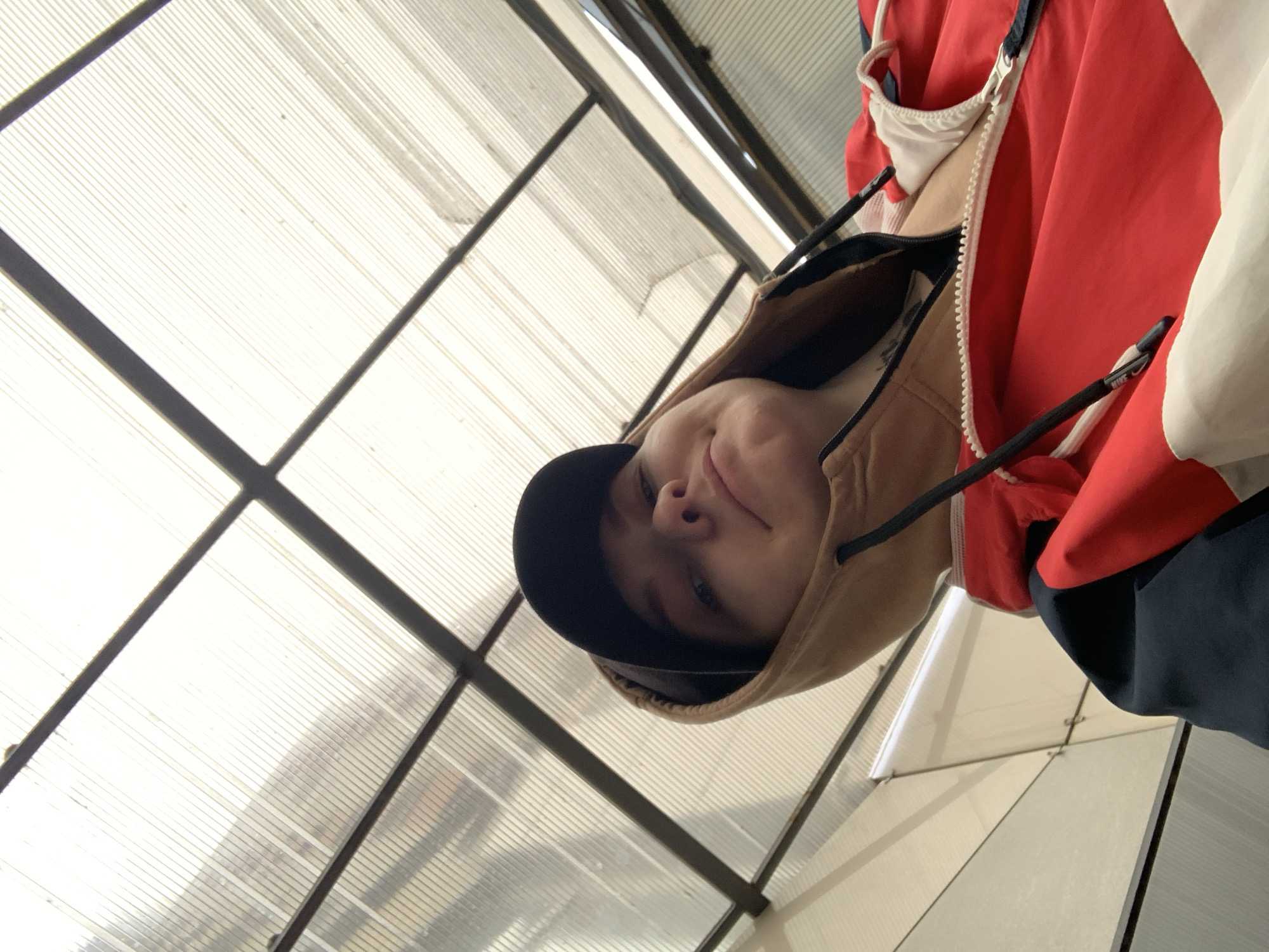 The image shows a person wearing a black cap and a tan hooded sweatshirt, with a red, white, and navy blue jacket partially visible. The person is looking down at the camera with a slight smile. The background features a translucent panel structure with black metal supports. The location is in the city of Unknown, Unknown.