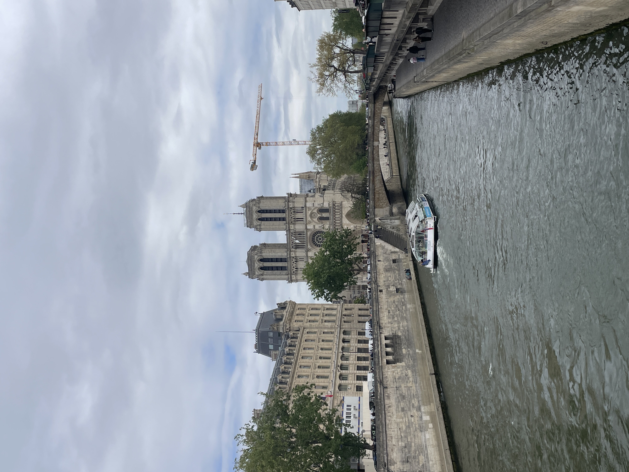 The image depicts the Seine River in Paris, France, with the Notre-Dame Cathedral prominently featured. The cathedral is under construction, evident by the presence of a large yellow crane behind it. A tour boat filled with people navigates the river. Stone embankments line the riverbanks. Trees grow along the embankment, and some people are walking. The sky is overcast with grey clouds. Buildings are visible on both sides of the river.