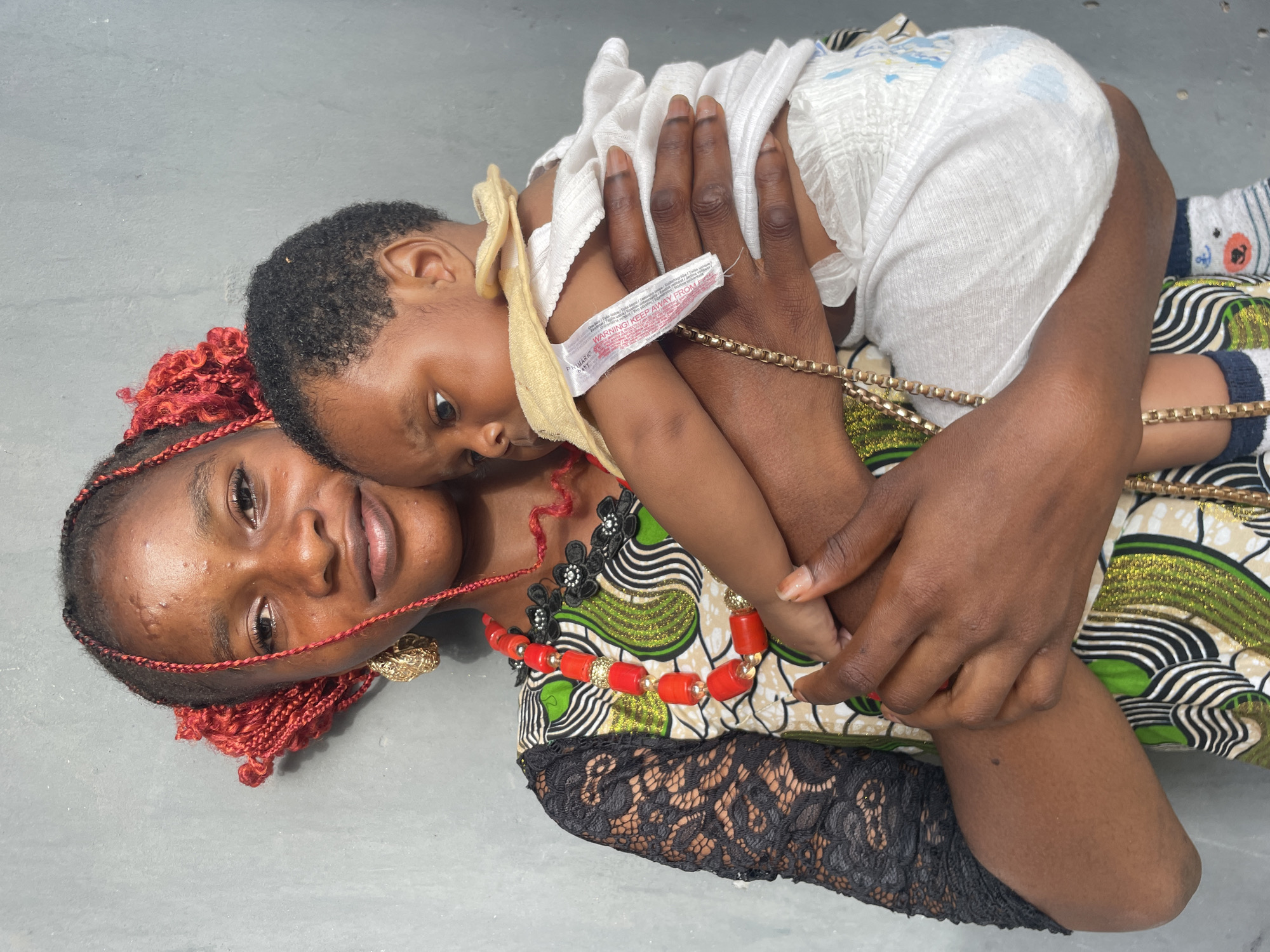 An intimate portrait shot from above captures a woman and a baby lying down, with the woman holding the child close. The woman, with dark skin, looks directly at the viewer with a slight smile, suggesting contentment. Her hair is styled in red braids, with some individual red and black braids framing her face. She wears multiple necklaces, including a prominent red and gold beaded one and another black and white beaded one, along with large gold earrings. Her dress features a vibrant African print in shades of green, white, black, and brown, with black lace sleeves.
The baby, also with dark skin and short dark curly hair, is nestled against the woman's chest and shoulder, looking towards the right. The baby is dressed in a white top and a white disposable diaper, with a yellow fabric item (possibly a burp cloth or part of an outfit) draped around its neck and chest. Visible on a white tag attached to this yellow fabric are the words "WARNING! KEEP AWAY FROM FIRE" and other smaller text, including what appears to be "PRIMARK". The baby also wears patterned socks with anchors and what looks like cartoon animal faces.
The woman's right arm is wrapped around the baby, and a gold-colored chain or bracelet is visible on her wrist. The background is a plain, light grey surface, likely a concrete floor, suggesting an indoor or shaded outdoor setting in Badagry, Nigeria. The overall mood of the image is tender and calm, highlighting the bond between the two individuals.