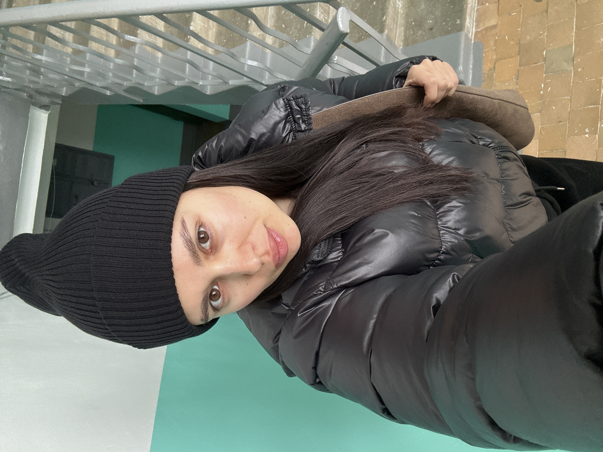 A woman is taking a selfie. She is wearing a black beanie and a shiny black puffer jacket. She has long, dark hair. She is holding a brown purse or bag in her hand. The background includes a metal railing, a wall painted in shades of green and white, and a brick wall. The location is likely in Chaikovsky, Russia, based on the request parameters.
