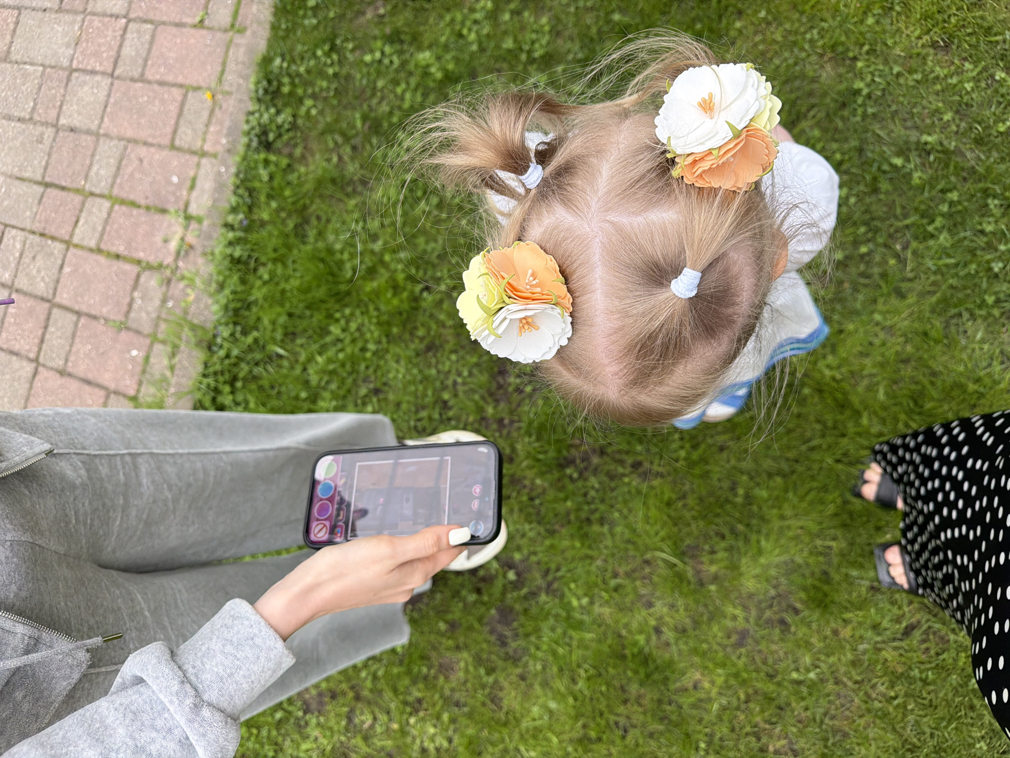 A top-down perspective captures a scene in a grassy outdoor setting, potentially a park or garden. A young child with fair hair styled in pigtails adorned with large floral hair accessories is the central subject. A woman's legs, clad in gray sweatpants and a matching sweatshirt, occupy the left side of the frame. Her hand holds a smartphone, its screen displaying an image that appears to be of an interior room. To the right of the child, a portion of another person is visible, wearing a black and white polka dot patterned dress and black sandals. The background consists mainly of green grass with a section of paving stones visible at the top left of the image. It's not possible to determine that the location is Moscow, Russia, from the image provided.