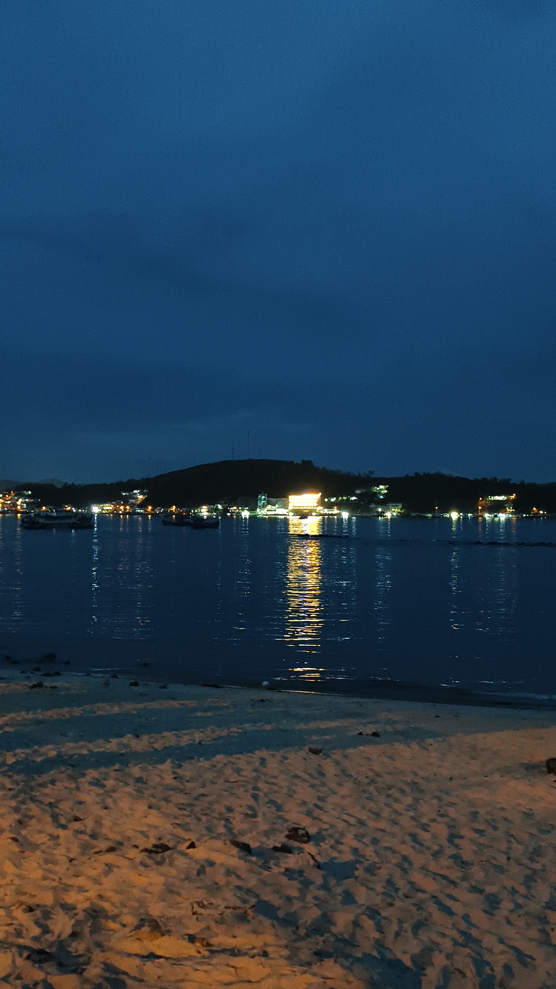 A nighttime view of a coastal city in Nha Trang, Vietnam, shows a body of water reflecting the lights of buildings and boats along the shoreline. The foreground features a sandy beach with shadows cast across it. The sky is dark blue and cloudy, indicating twilight or nighttime. Lights from buildings and boats are visible along the distant hilly coastline.