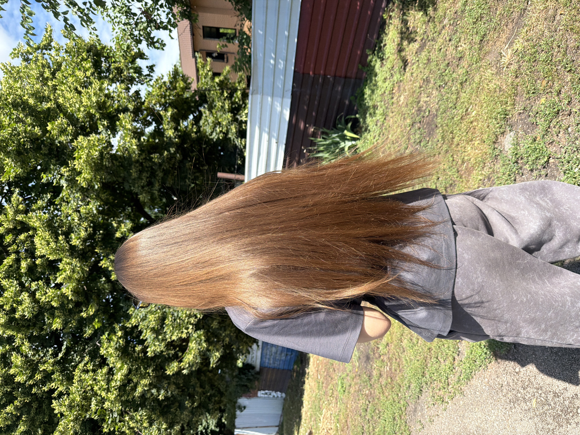 A person with long, reddish-brown hair is standing outside, viewed from the back, facing away from the camera. They are wearing a dark grey t-shirt and grey sweatpants. The person is in an outdoor environment with green grass and trees. In the background, there is a fence with white and brown sections, and a building with a brown roof. The scene appears to be in a residential area, possibly a backyard or a street in the city of Dnipro, Ukraine, given the architectural style of the building and the type of fencing. The lighting suggests it is daytime, with bright sunlight casting shadows on the ground.