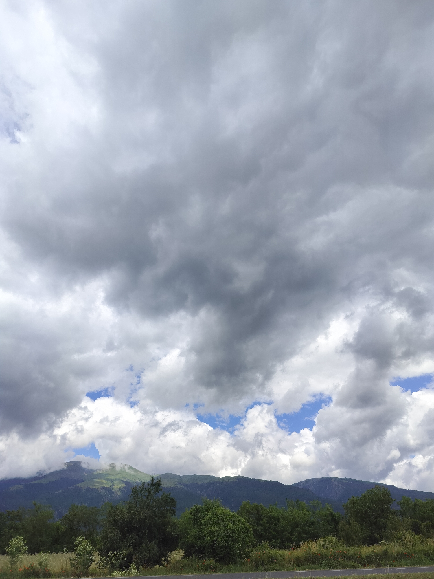 The image depicts a cloudy sky over a mountainous landscape in Tarnicheni, Bulgaria. The foreground features a line of trees and vegetation, with a road visible at the bottom. The middle ground is occupied by rolling hills and mountains, some of which are shrouded in mist or clouds. The sky is filled with a mix of dark, heavy clouds and patches of blue sky, suggesting a dynamic weather system. No people or specific actions are visible in the image. The overall scene conveys a natural, somewhat dramatic, outdoor environment.