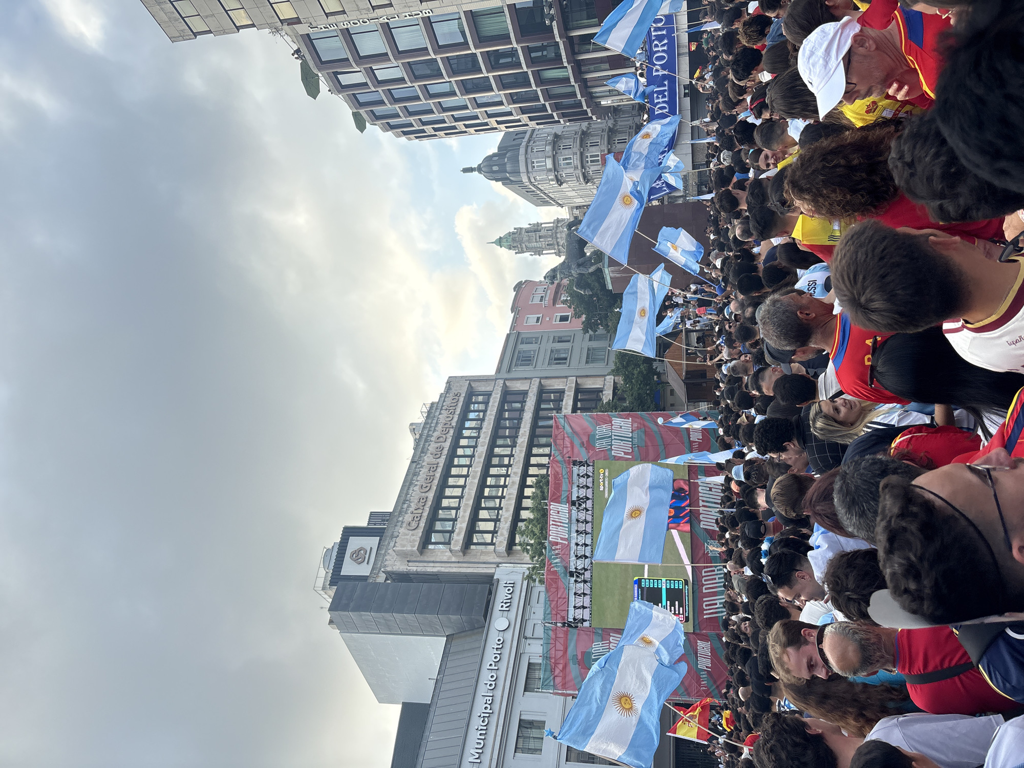 The image depicts a densely packed crowd of people in an urban setting, likely in Porto, Portugal, given the flags and banners. Many individuals are waving or holding numerous light blue and white striped flags with a sun emblem, which are the flags of Argentina. Some banners in the background also feature text that appears to be in Portuguese, including "MUNICIPAL DO PORTO RIVOLI" and "CAIXA GERAL DE DEPOSITOS," indicating a location in Porto. There are large screens in the background displaying what appears to be a sports broadcast, possibly a football match, with scores and player names visible. The crowd is comprised of people of various ages and ethnicities, predominantly dressed in casual clothing, with some wearing football jerseys in team colors. The environment includes modern and older architectural buildings, some with large windows and signage. The sky is overcast. The overall atmosphere suggests a public viewing event or celebration related to a sporting event, with a strong presence of Argentinian supporters.