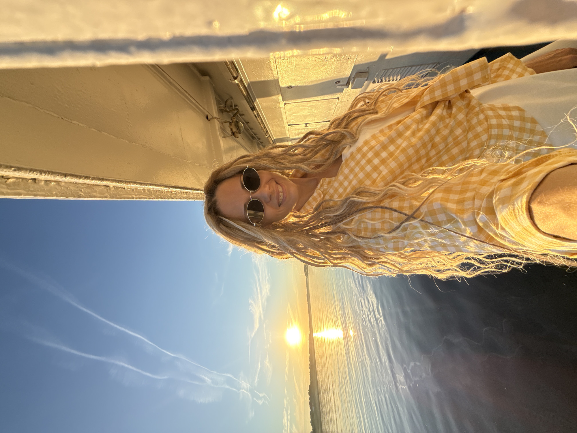 This image depicts an adult female positioned on the exterior of a large vessel, likely a ship or boat, at an unspecified location within Rusanovo, Russia, during golden hour (either sunrise or sunset).
The female subject, with medium skin tone, is smiling broadly at the camera, revealing teeth. She wears dark, oval-lensed sunglasses with silver frames. Her long, light blonde hair is styled in a combination of dreadlocks and wavy strands, cascading over her shoulders. She is dressed in a yellow and white gingham (checked) long-sleeved shirt, partially unbuttoned over a plain white garment, possibly a tank top or dress. The sleeves of her shirt are rolled up. Her right arm, visible in the lower right foreground, appears to be holding a device, likely the camera for a selfie, or is positioned close to the camera.
To the subject's right (viewer's left) and behind her, the sturdy, off-white or cream-colored painted metal structure of the vessel is visible, heavily illuminated by the golden light. This structure includes what appear to be bolted panels, pipes, and some fixtures. The underside of an upper deck or overhang is also visible directly above the subject.
The background features a vast body of water, likely a river or lake, stretching towards the horizon. The sun, appearing as a bright, circular disc, is positioned low on the horizon over the water, casting a golden reflection path directly towards the viewer. The water surface shows gentle ripples and subtle dark patterns. A distant landmass, possibly a shoreline with trees and faint structures, is visible on the horizon beneath the sun.
The sky is a clear, light blue, transitioning to warmer tones near the horizon. Two prominent, parallel white contrails (condensation trails from aircraft) arc across the upper left quadrant of the sky, with a fainter third trail also discernible. The overall lighting is warm and bright, originating from the low sun, creating strong highlights and shadows across the scene.