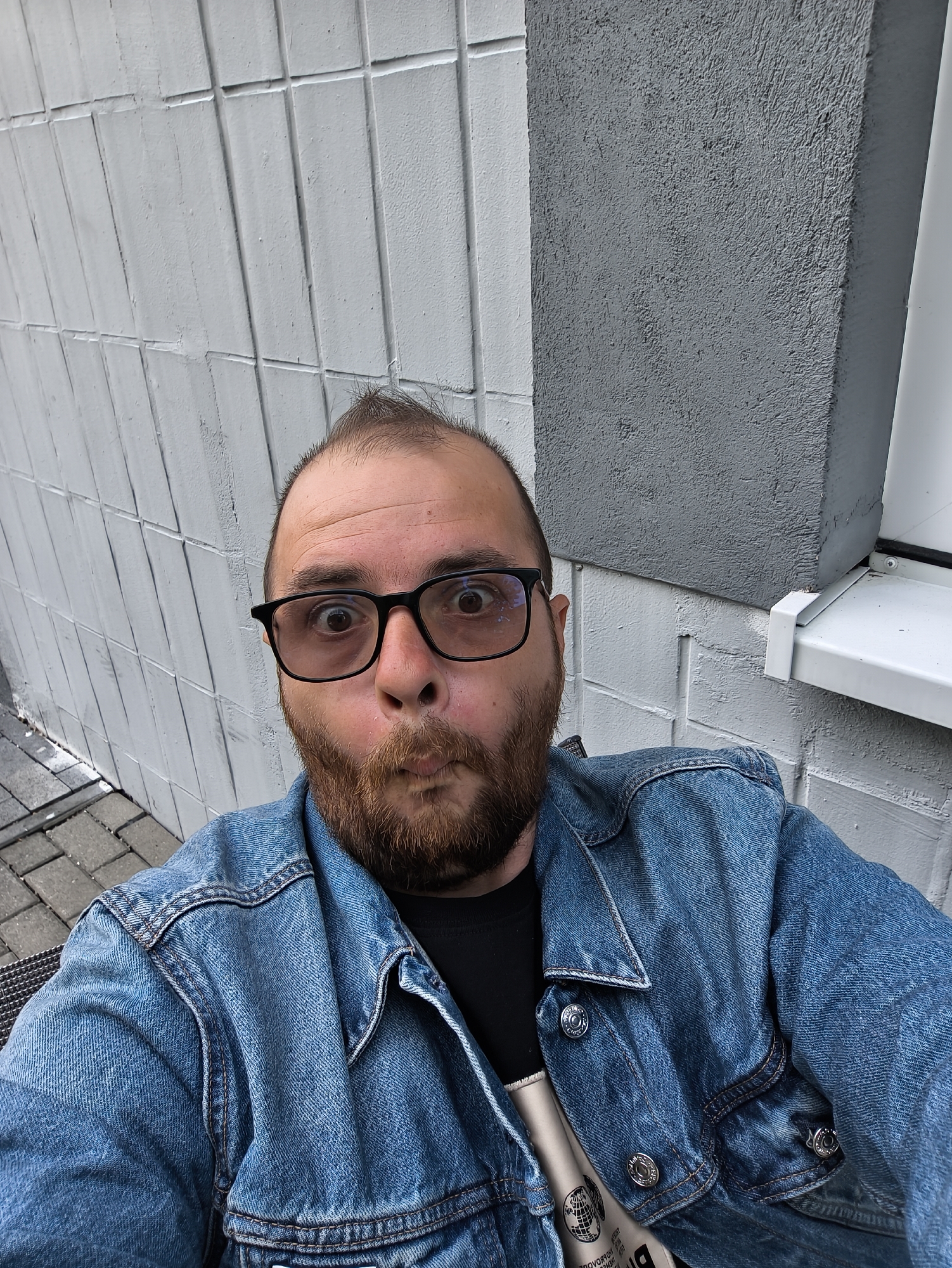 A man with a beard and glasses is taking a selfie. He is wearing a denim jacket over a black shirt. He is making a surprised face with his mouth puckered. The background is a gray and white brick wall and a window sill. The location is an urban setting, possibly in the city of Unknown, Unknown.