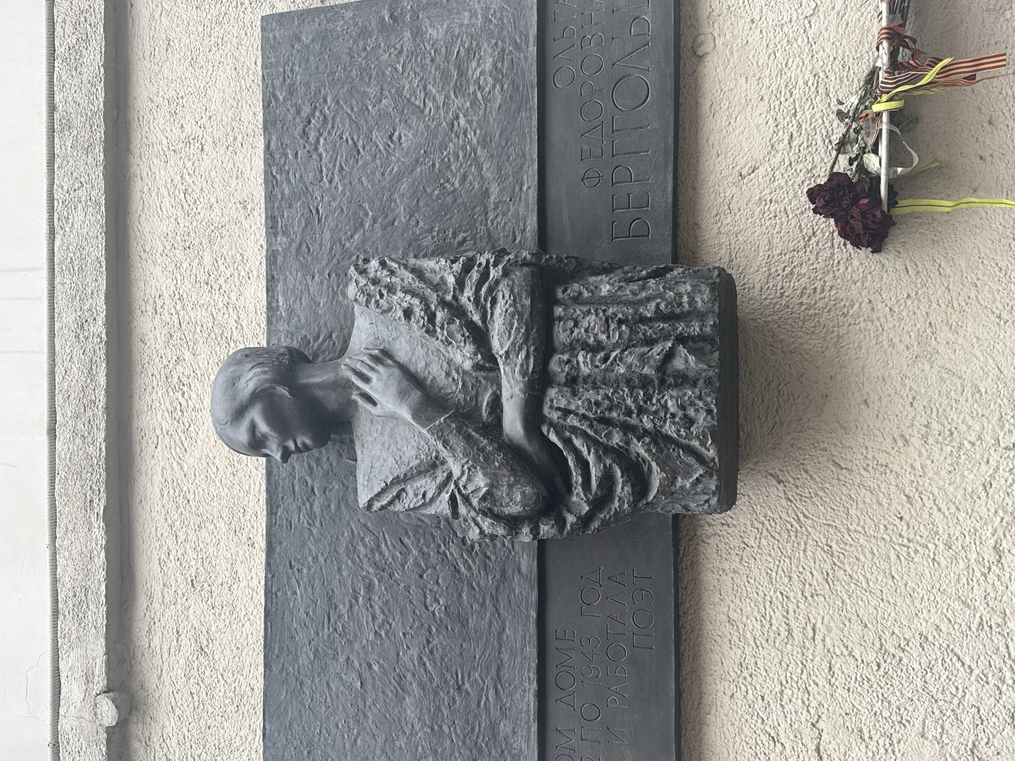 The image shows a memorial plaque on a light beige, textured wall. The plaque is vertically oriented and dark grey. On the right side of the plaque, the inscription "ОЛЬГА ФЕДОРОВНА БЕРГГОЛЬЦ" (Olga Fedorovna Bergholz) is visible in Cyrillic. The central part of the plaque features a three-dimensional sculpture of a person's head and upper body in relief, with a hand resting on their chest. Below this relief, another inscription in Cyrillic is visible, including the years "1942-1943" and the word "ПОЭТ" (poet). To the right of the plaque and slightly above it, a small bunch of faded flowers is attached to the wall. The flowers are mixed with ribbons, one of which has the colors of the Ribbon of Saint George (orange and black). The location is likely Saint Petersburg, Russia, given the Cyrillic inscription and the cultural significance of Olga Bergholz.