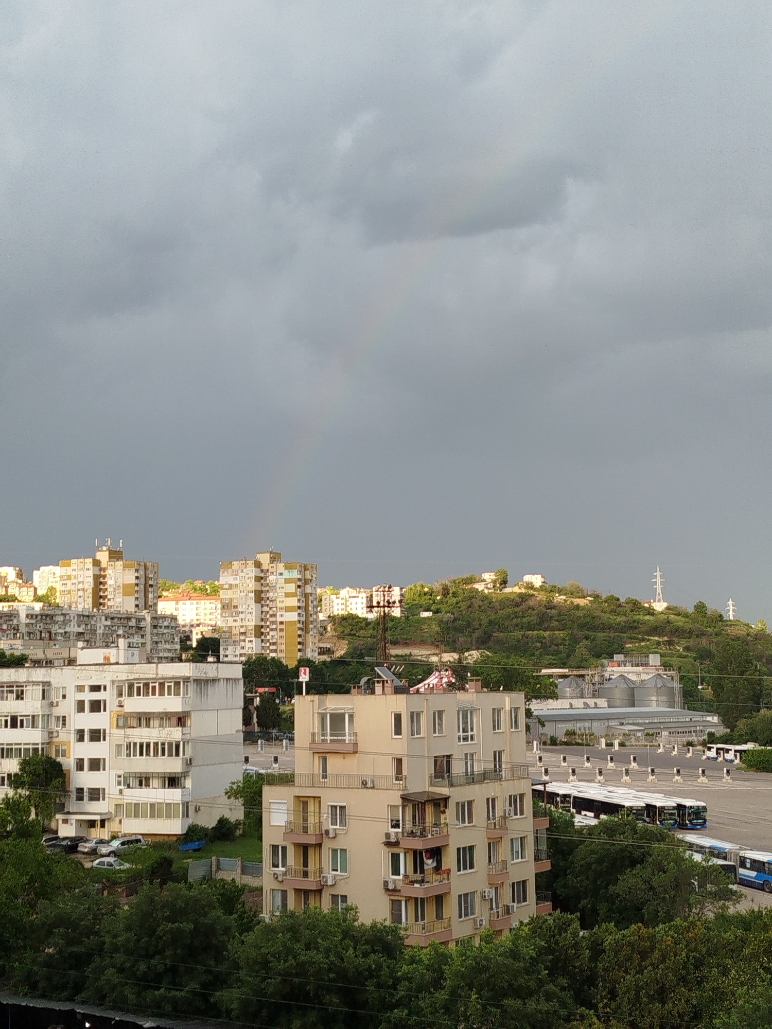 The image shows a cityscape under a cloudy sky with a faint rainbow visible. It appears to be taken in Varna, Bulgaria, overlooking a dense urban area with numerous apartment buildings of varying heights and architectural styles. In the foreground, trees obscure the lower portion of the buildings.
To the right, there is a bus parking lot with several buses lined up. Beyond the buildings, a hill rises, covered with greenery and some structures, including what appears to be a transmission tower. The sky is overcast and gloomy, but the presence of the rainbow suggests a recent or ongoing rain shower.