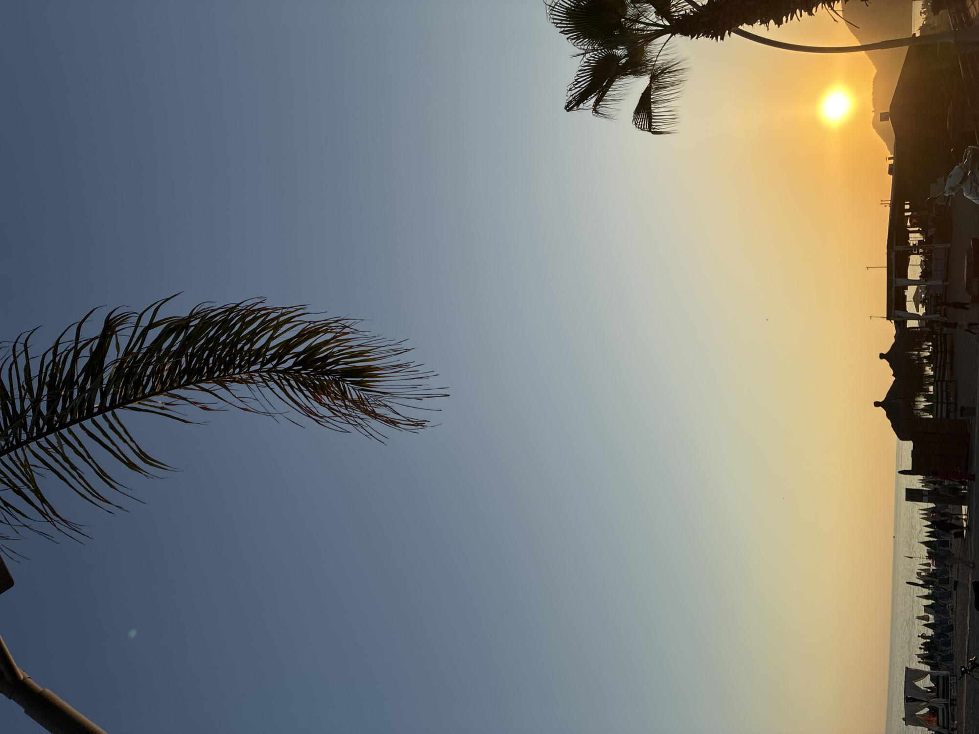 The image captures a serene sunset scene at a beach in Alanya, Turkey. The sky transitions from a clear blue at the top to a warm orange hue near the horizon. Silhouetted against the setting sun are palm trees, their fronds delicately framing the sky.
In the foreground, the beach is dotted with beach chairs, umbrellas, and several thatched-roof structures, likely beachfront cafes or bars. The sea is visible in the distance, with a calm surface reflecting the sunset colors. A few people are scattered across the beach, some appearing to be walking or relaxing. The overall atmosphere is tranquil, characteristic of a peaceful evening on the Mediterranean coast. The silhouette of a mountain range can be seen in the far distance, further enhancing the natural beauty of the scene.
