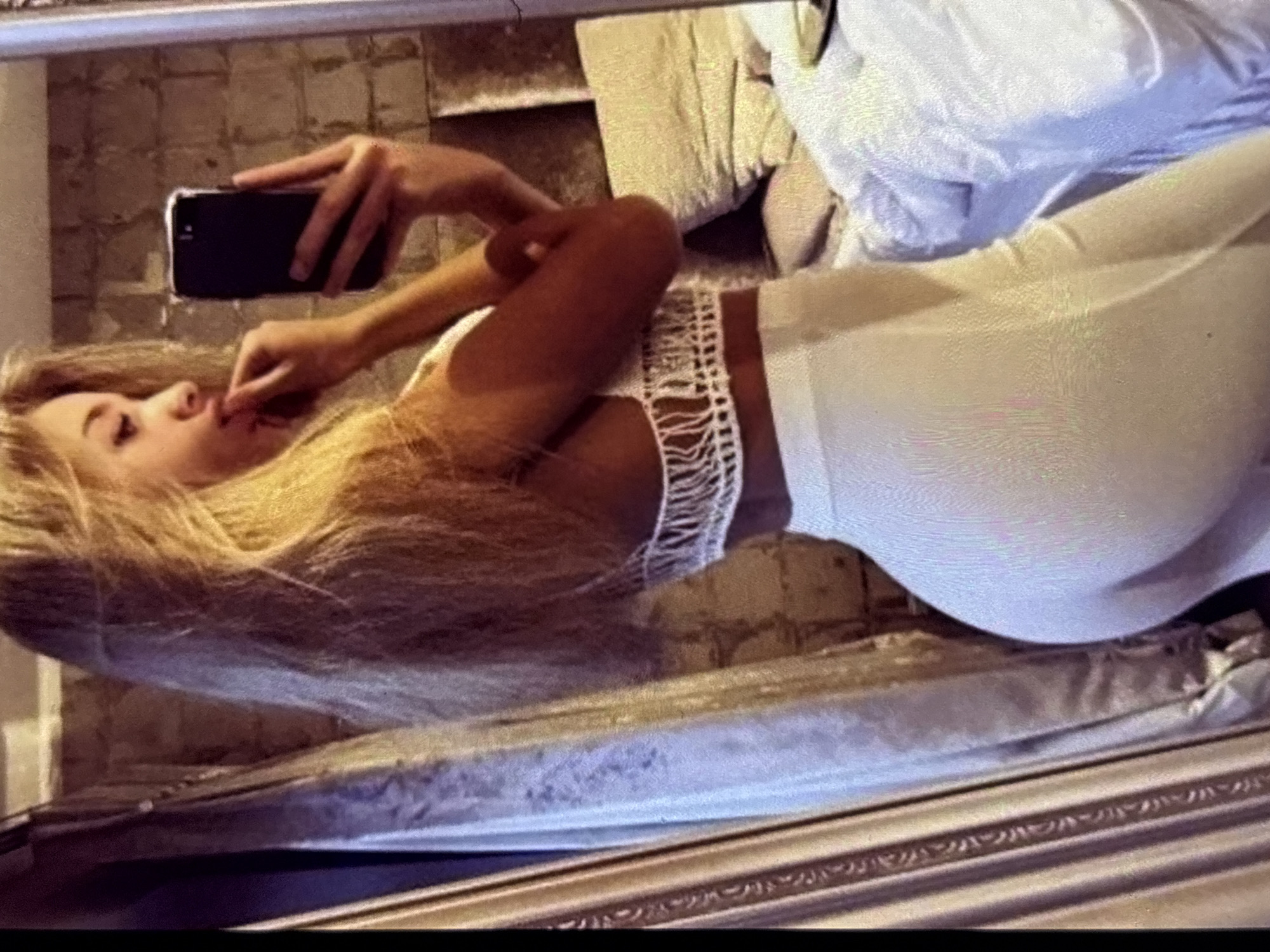 A young woman with blonde hair is taking a selfie in a room. She is wearing a white crochet halter top and white pants. She is standing in front of a mirror, which reflects her and the room. The room appears to be a bedroom, with a bed and curtains visible in the background. The woman is touching her lips with her fingers.