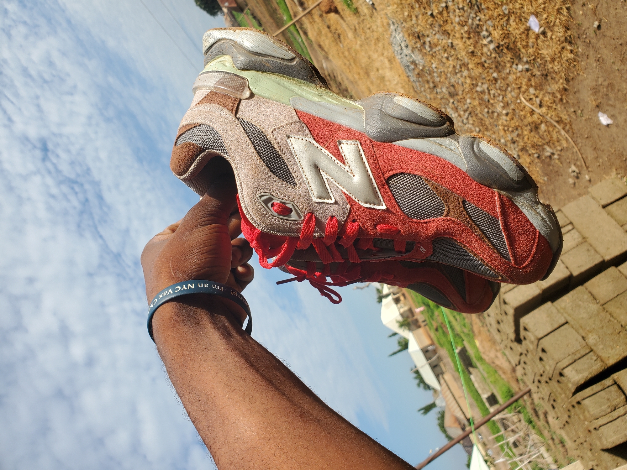An outdoor, eye-level shot captures a dark-skinned arm and hand holding a pair of New Balance sneakers against a bright sky in the city of Winters, United States of America. The hand, positioned in the upper center, firmly grips the shoes. A blue wristband on the forearm features white text, partially legible as "NYC Vax Champ!". The sneakers are predominantly red suede and dark gray mesh with light beige/tan suede overlays, showcasing a distinct light gray "N" logo on the side and red laces. The complex sole of the shoe is multi-toned in light green, dark gray, and light gray, with visible dirt and scuff marks. The background features a clear blue sky with scattered white clouds, suggesting a sunny day. Below the horizon, a dry, dusty landscape stretches out, dotted with low-rise buildings with light roofs and patches of green vegetation. In the lower left foreground, stacks of gray concrete blocks are visible on the dry, light brown earth.