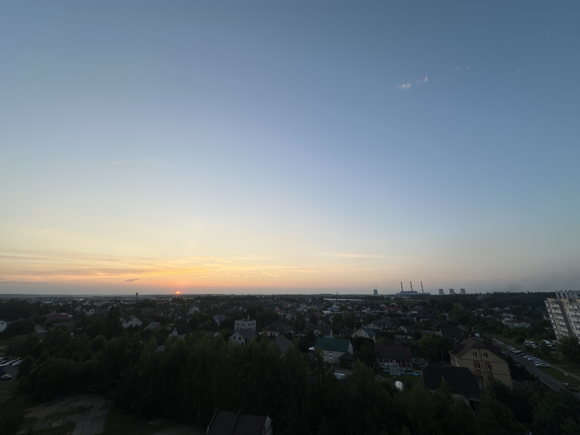 The image depicts a wide panoramic view of a suburban landscape at dusk, with the sun setting on the horizon. In the foreground, dense green trees dominate the lower portion of the frame, obscuring some of the residential buildings. The middle ground reveals a sprawling residential area with numerous houses, varying in architectural style and color, interspersed with more trees and some paved roads. In the distance, industrial structures, specifically cooling towers of a power plant, are visible against the fading light of the sky. The sky transitions from a warm orange and yellow hue near the horizon, indicative of the sunset, to a clear, pale blue overhead. A few wispy clouds are scattered across the upper sky. The scene appears to be set in Шчомысліцкі сельскі Савет, Belarus, given the architectural styles and the presence of the industrial facility which is characteristic of the region. There are no people visible in the image. The overall impression is one of a quiet, developing suburban area on the edge of industrial activity.