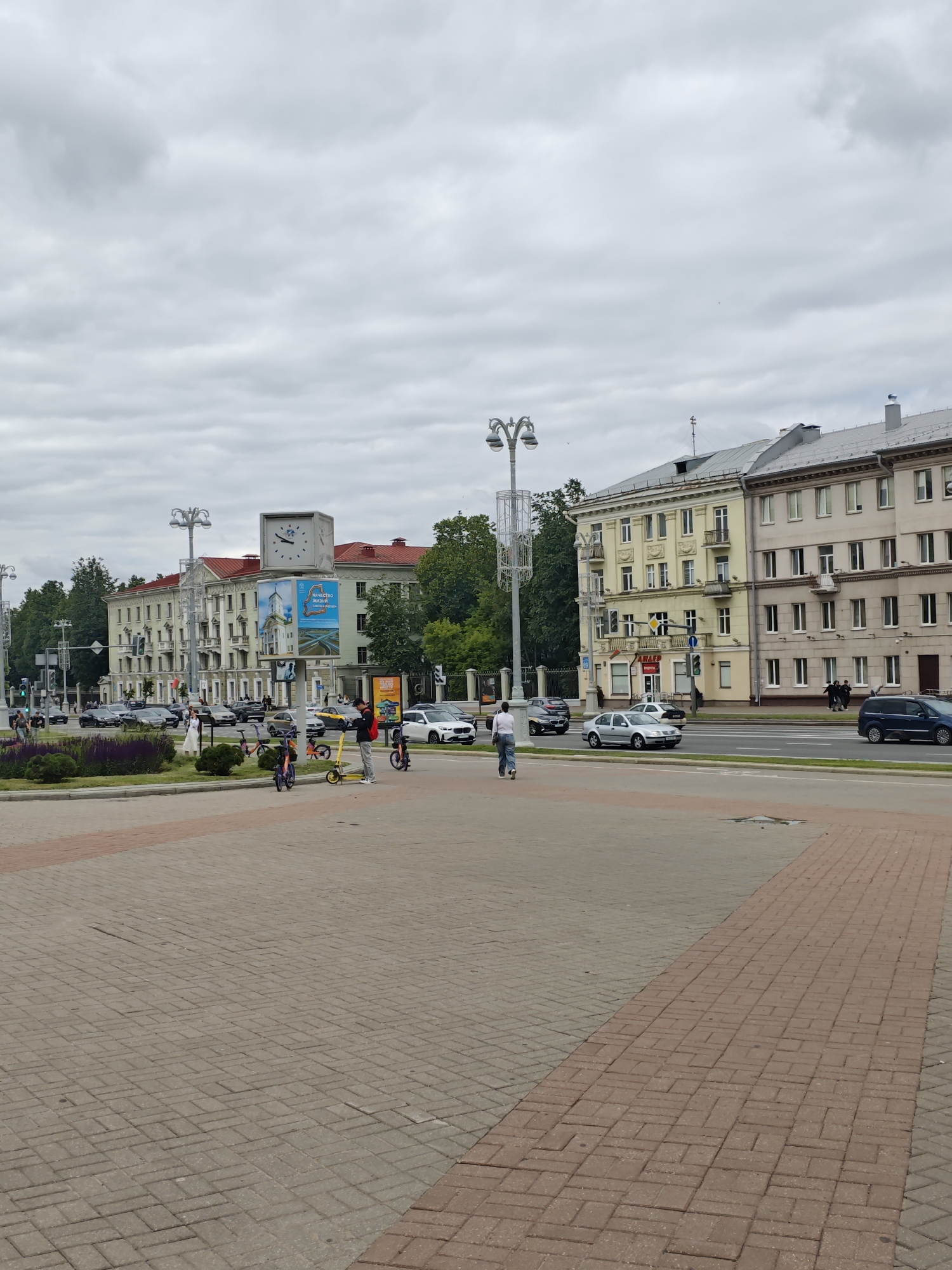 The image shows a street scene in Minsk, Belarus, with buildings lining the left side of the street and a large plaza in the foreground. The buildings are multi-story structures with red roofs and light-colored facades. A large clock is visible on top of one of the buildings. Several cars and people are on the street, and there are some electric scooters parked on the plaza. The sky is overcast. The plaza is paved with brick pavers, with a pattern of red and gray bricks. A red brick path runs diagonally across the foreground. The scene appears to be a public space in a city center.