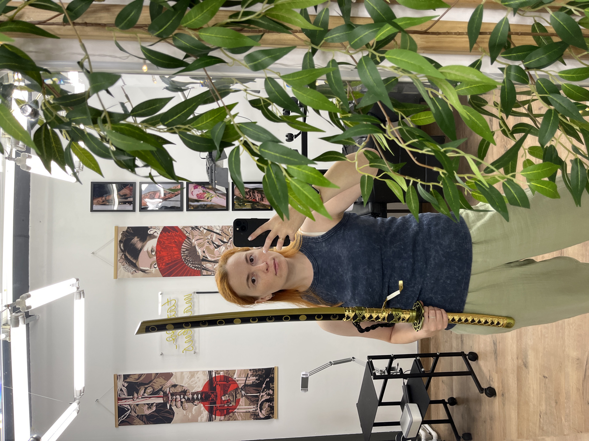 A red-haired woman stands in a well-lit room, possibly a tattoo studio, holding a decorative sword with a gold-colored hilt and sheath. She is wearing a dark blue tank top and light green pants, and is taking a selfie with her phone. The room features several pieces of Japanese-inspired artwork hanging on the wall, along with a neon sign. A large artificial plant with green leaves obscures part of the right side of the image. The floor is made of wood planks, and there is a trolley with equipment in the background. The presence of tattoo-themed art suggests this location could be in a city known for such establishments, though its specific location in Chaikovsky, Russia, cannot be determined from the visual information provided.