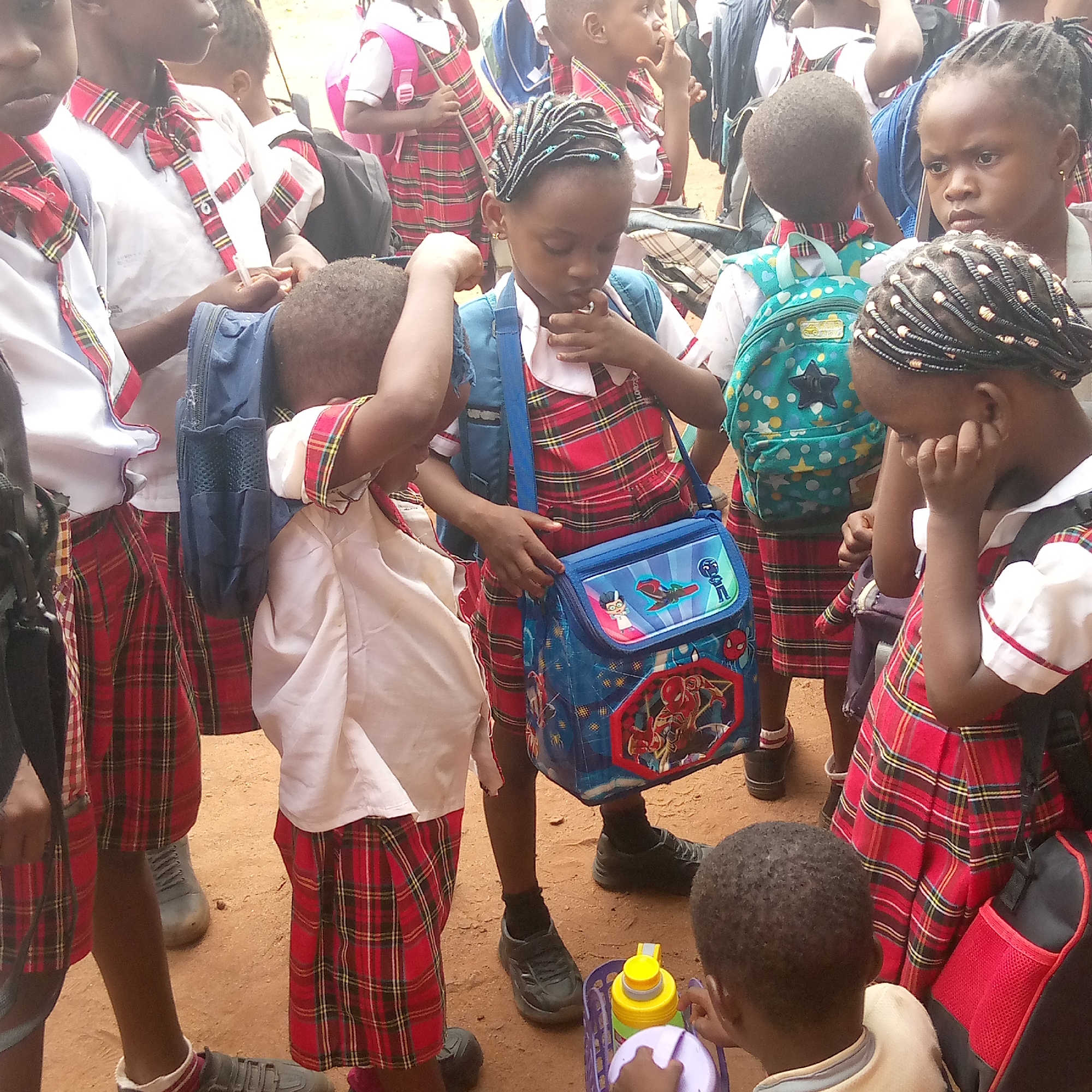 The image shows a group of African children wearing red and white plaid school uniforms. They are standing on what appears to be a reddish-brown dirt ground. Some children are adjusting their backpacks, while others are holding lunch bags. One child is wearing a blue lunch bag with superhero images on it. The image captures a school gathering in Ukwu-Abwa, Nigeria.