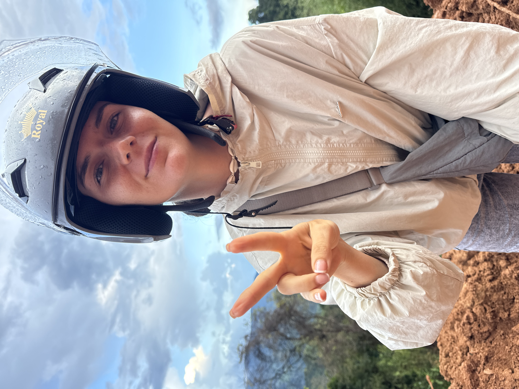 In this close-up selfie, a young woman wearing a dark grey helmet with a clear visor and a golden emblem that says "Royal" is pictured outdoors. She is looking directly at the camera with a faint smile, her right hand raised in a peace sign. She is wearing a light beige hooded jacket with a zipper and a grey strap bag slung across her body. The background shows a cloudy sky with patches of blue and hints of greenery from trees and foliage, suggesting a natural or rural setting, possibly in or around Nao Sẻ, Vietnam. The terrain in the foreground appears to be made of reddish-brown soil. The helmet has visible water droplets on it, indicating recent rain or high humidity.
