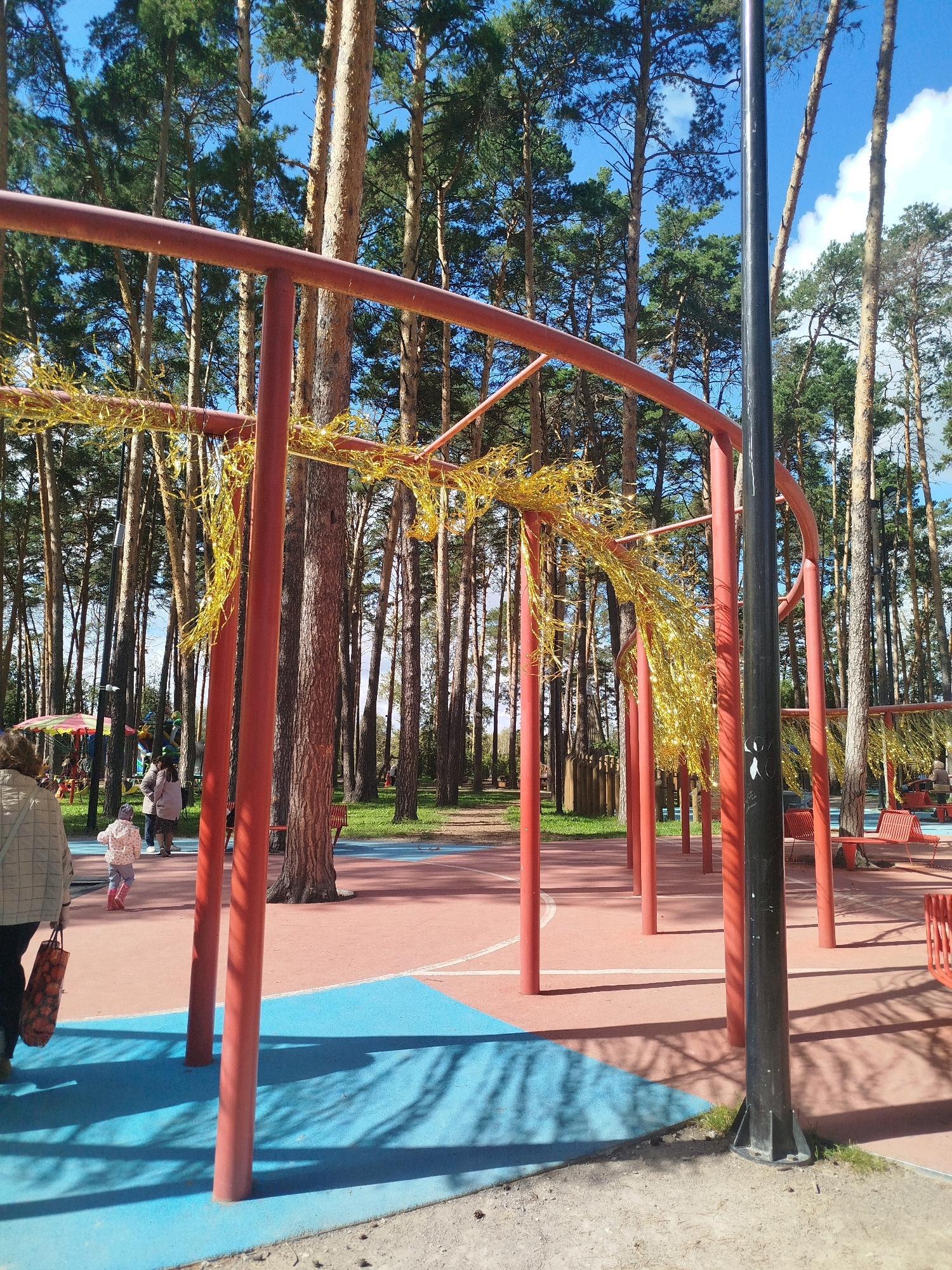 The image depicts an outdoor recreational area within a pine forest in Zavodoukovsk, Russia. A large, curved red metal structure, possibly part of a playground or sports equipment, dominates the foreground and midground. Golden tinsel-like decorations are draped across the structure. A black lamppost stands to the right of the red structure. The ground is a paved surface with sections of pink and blue, indicating a sports court or play area. In the background, numerous tall pine trees create a dense forest. On the left side of the image, a woman in a checkered jacket and dark pants is walking away from the camera, carrying a patterned bag. Further back, a child in a light-colored jacket and pink boots runs on the blue section of the ground. Several other people are visible in the midground, some standing and interacting. Red benches are present in the background, and a colorful umbrella-like structure, likely part of a playground, is also visible. The sky is blue with some white clouds. The overall scene suggests a public park or playground on a sunny day.