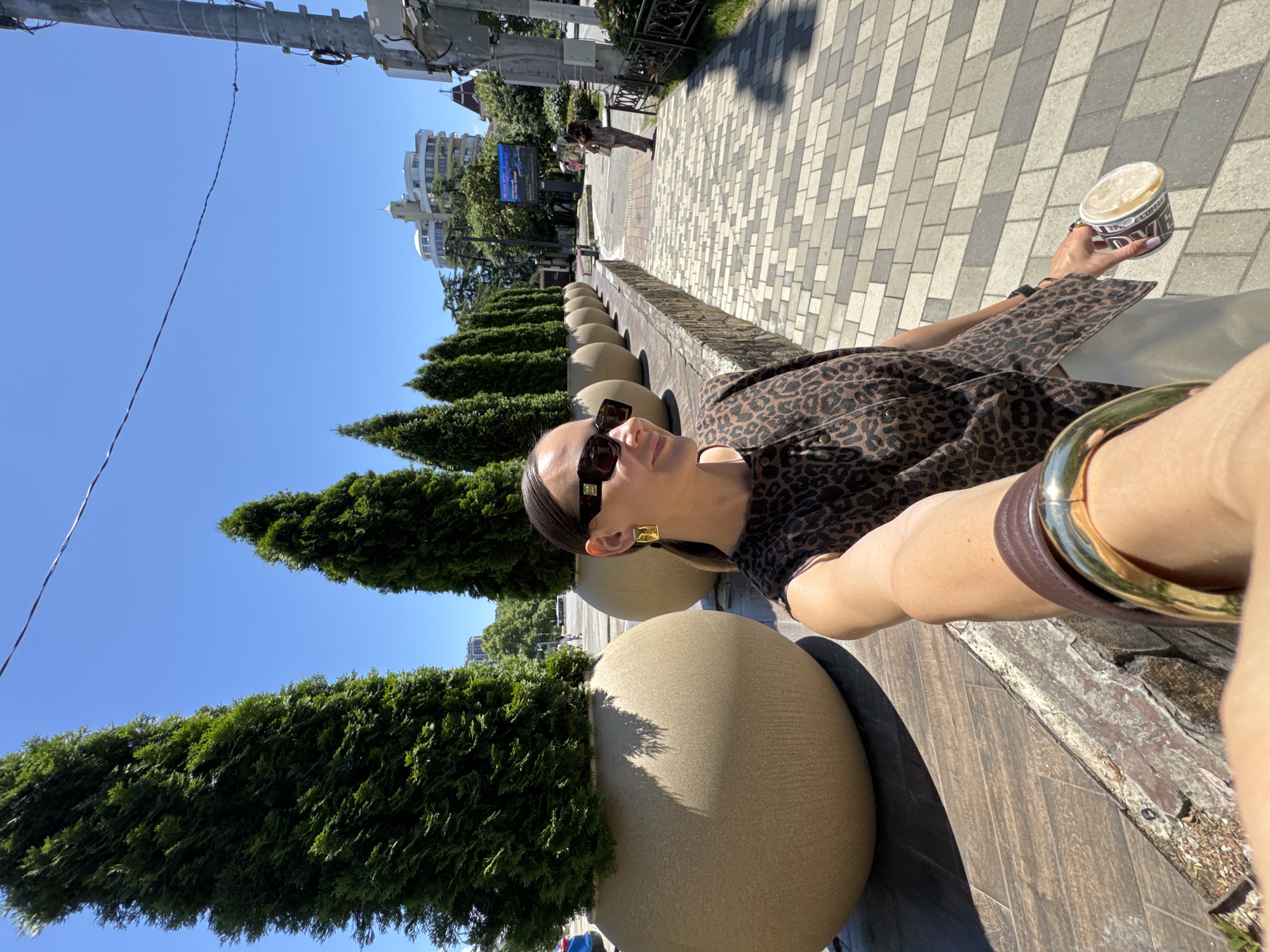 In this selfie taken outdoors on a sunny day in Sochi, Russia, a woman with dark hair pulled back and wearing large, rectangular, dark sunglasses and a leopard-print top is holding a coffee cup in her right hand and extending her left arm to capture the shot. She is wearing a gold, square-shaped earring and a thick gold bracelet on her left wrist. She is standing on a paved walkway next to a raised planter filled with a tall, dark green conifer tree. Behind her, a line of similar planters with trees stretches into the distance. The walkway is made of light-colored rectangular pavers arranged in a pattern. In the background, buildings, trees, and a utility pole are visible. A second woman in a patterned outfit is walking away in the distance on the right side of the frame. The overall environment suggests a well-maintained urban public space, possibly a promenade or park area.