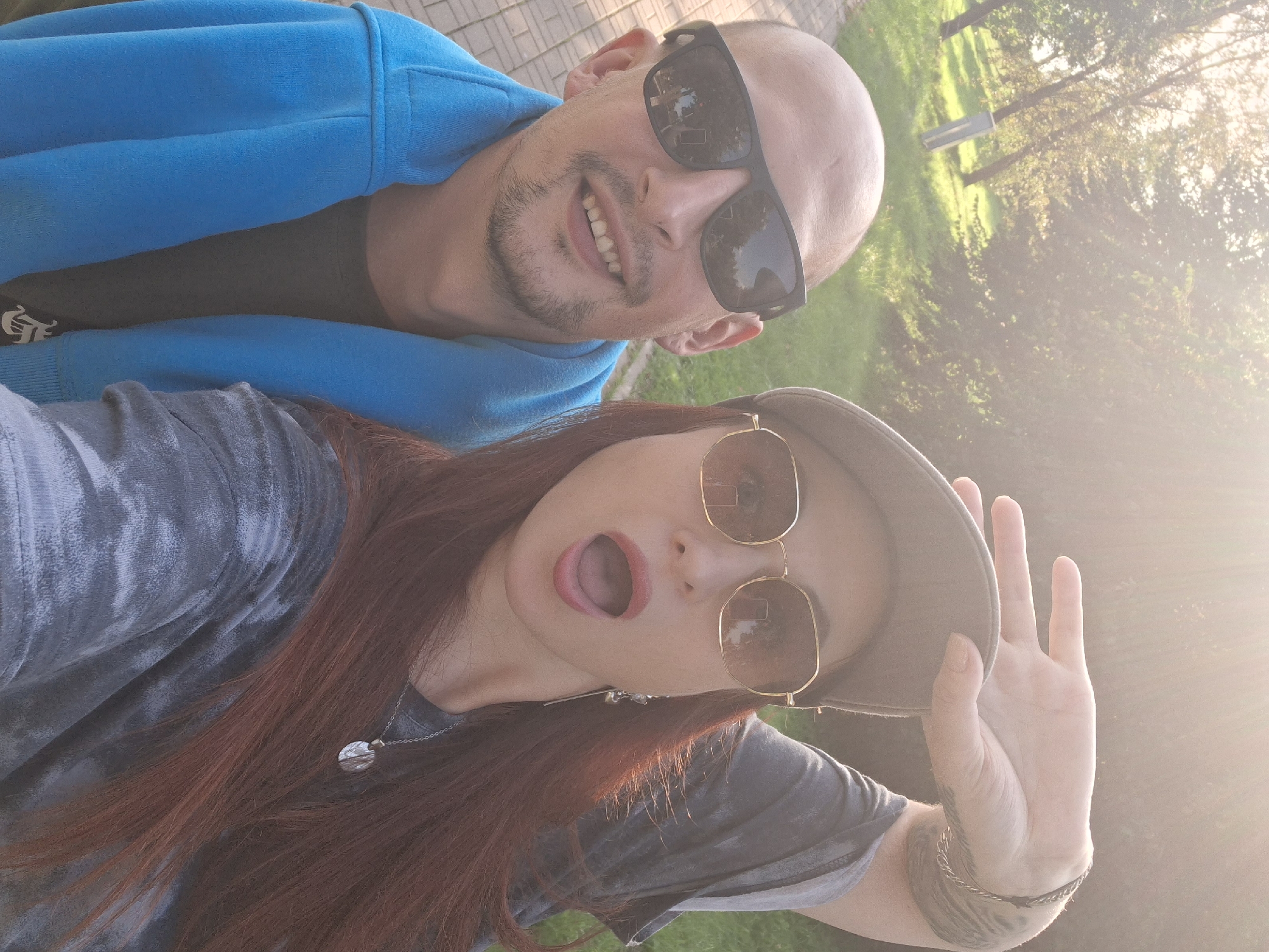 A man with a shaved head and sunglasses smiles at the camera. He is wearing a bright blue hooded sweatshirt over a dark t-shirt. To his right, a woman with long auburn hair, wearing a baseball cap and sunglasses, makes a peace sign with her hand. Her mouth is open, and she is wearing a tie-dye shirt and a necklace with a circular pendant. They are both outdoors, with trees and grass in the background. The lighting suggests it is daytime, with the sun casting a warm glow. The location is the city of Myadzyel, Belarus.