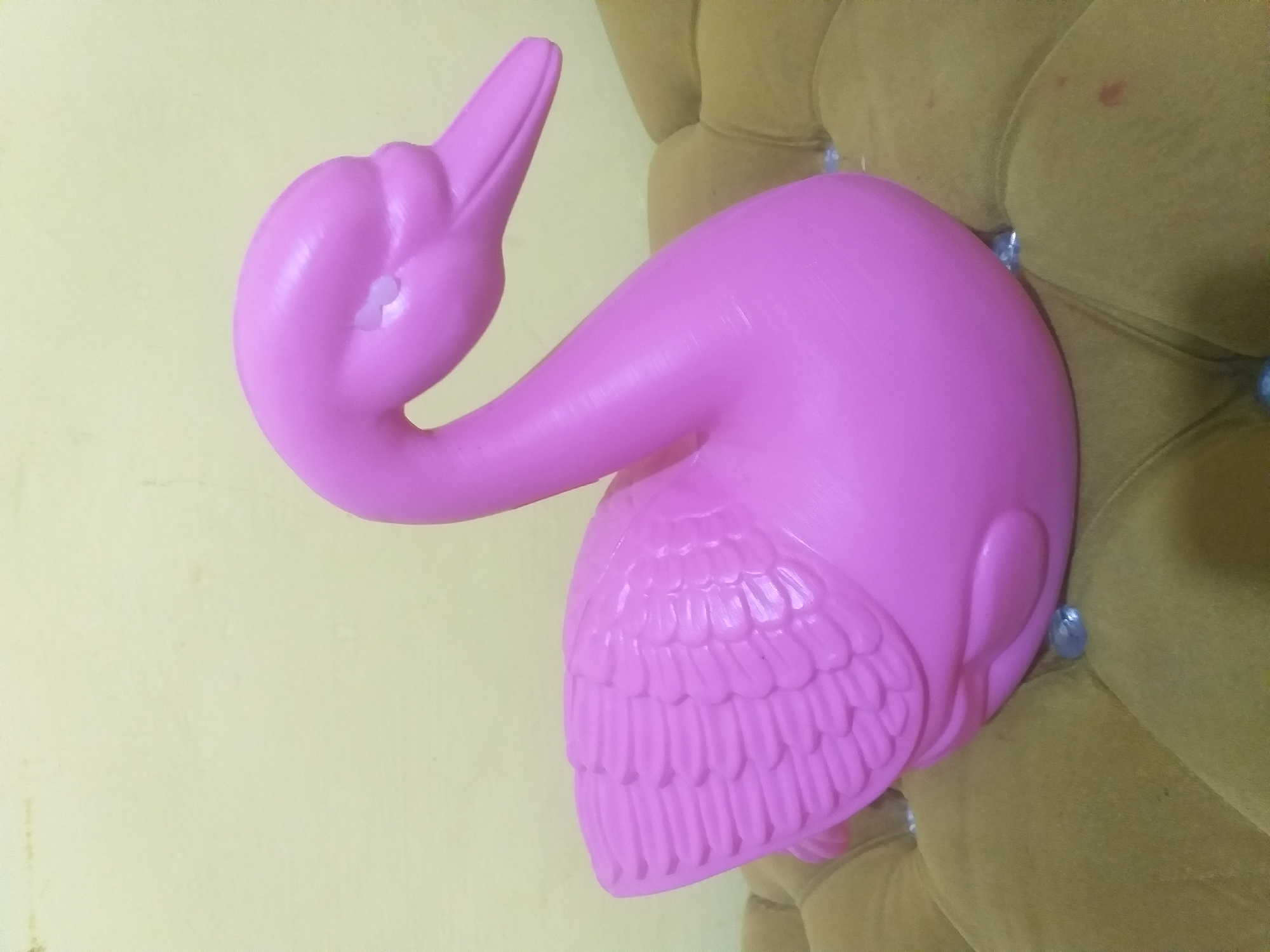 A pink plastic swan figurine is positioned on a tufted brown sofa. The swan is facing right, with its head slightly tilted upwards. The background wall is a pale yellow color. There is no indication of the specific location within Dumai City, Indonesia.