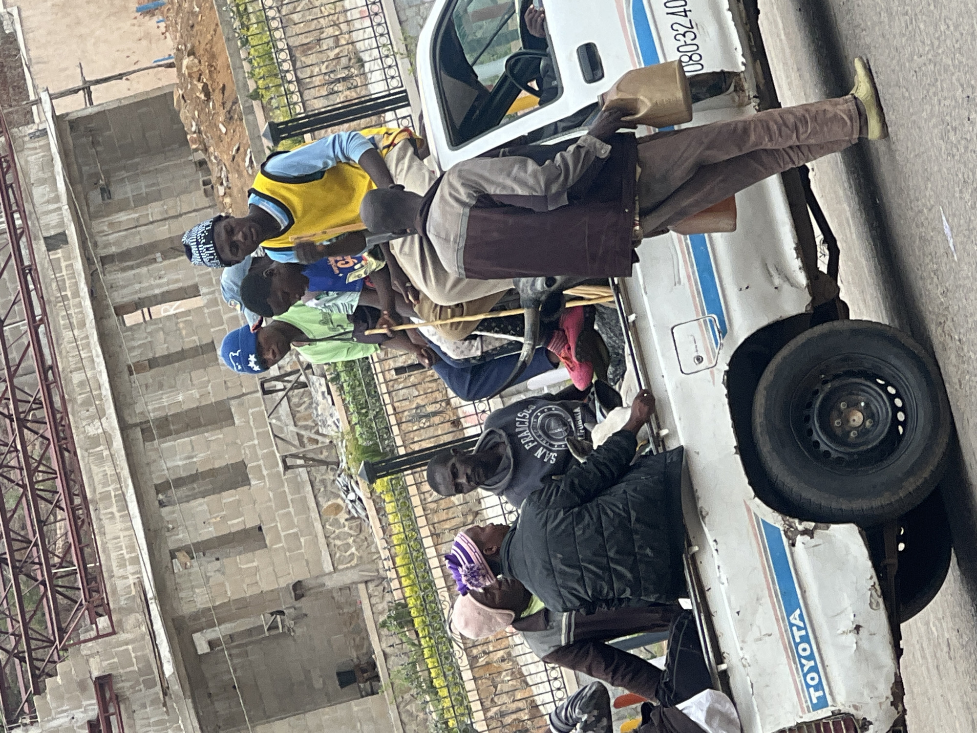 The image captures a daytime street scene in Jos, Nigeria, featuring a group of approximately 8-10 men and boys gathered around and within an old, white Toyota pick-up truck. Most individuals appear to be young Black men or older boys.
At the center, a man in a light brown jacket and trousers stands beside the truck, holding a yellow plastic jerrycan, potentially carrying fuel or water. Inside the truck's bed, several other people are visible, some sitting, others leaning. Another individual is seated in the driver's seat. The white Toyota pick-up shows considerable wear, with rust spots and peeling paint, and features distinctive blue and red stripes along its side. The word "TOYOTA" is printed in blue letters, and a partial phone number, "080-3240", is visible on the truck's exterior.
In the background, an unfinished concrete block building, possibly under construction, rises behind a metal fence. Exposed rebar or scaffolding is visible on its left side. The ground transitions from a paved road where the truck is to a dirt and rubble area behind the fence.
The bright, even lighting indicates it is daytime, and the casual clothing worn by the individuals (t-shirts, hats, and light jackets, including one grey hoodie with "SAN FRANCISCO" text) suggests a mild to warm climate. The scene depicts a casual gathering, possibly waiting for transport or involved in an informal activity related to the truck. No strong emotions are discernable on the faces of the people.
