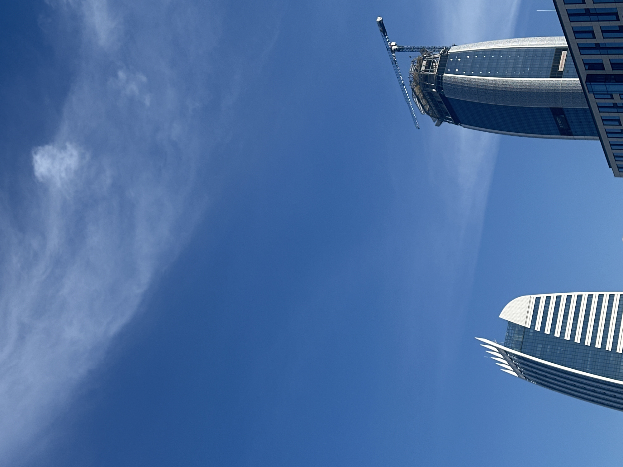 The image captures a striking low-angle perspective of a clear, vibrant blue sky dominating the frame, with a few delicate, wispy white clouds scattered across it. Two modern skyscrapers, characteristic of a contemporary urban environment, are partially visible along the right side of the composition.
The upper skyscraper, oriented almost horizontally, appears to be under construction or recently completed, with a prominent construction crane attached to its uppermost section. This building features a sleek, curved glass facade with sections of dark blue glass and metal paneling. Vertically visible on its side is the text "ETEM". A faint white airplane contrail can be seen high in the sky above this structure.
The lower building, also partially cropped and oriented horizontally, is fully constructed and showcases a distinct architectural design with a white, vertically ribbed or finned section at its peak, combined with glass surfaces.
The bright, unblemished blue sky and the crisp details of the buildings suggest it is daytime, likely a clear and sunny day. The scene conveys the modern architectural landscape of Sofia, Bulgaria, viewed from ground level looking upwards, emphasizing the height and design of the urban structures against the expanse of the sky. No people are visible in the image.