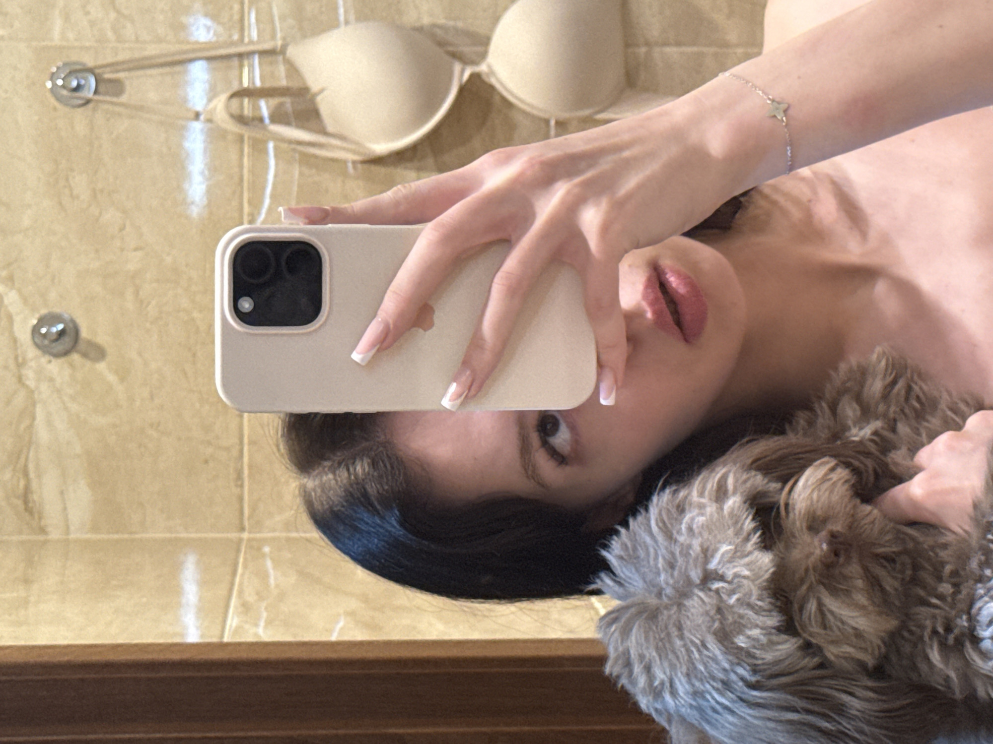 A young woman with dark hair and light skin takes a selfie in a bathroom, holding a small, fluffy brown dog. She is wearing a light-colored phone case and has manicured nails. A beige bra hangs on a hook behind her, and the walls are tiled with a marble pattern. The scene appears to be in an indoor setting, likely a bathroom in an apartment in Khimki, Russia.