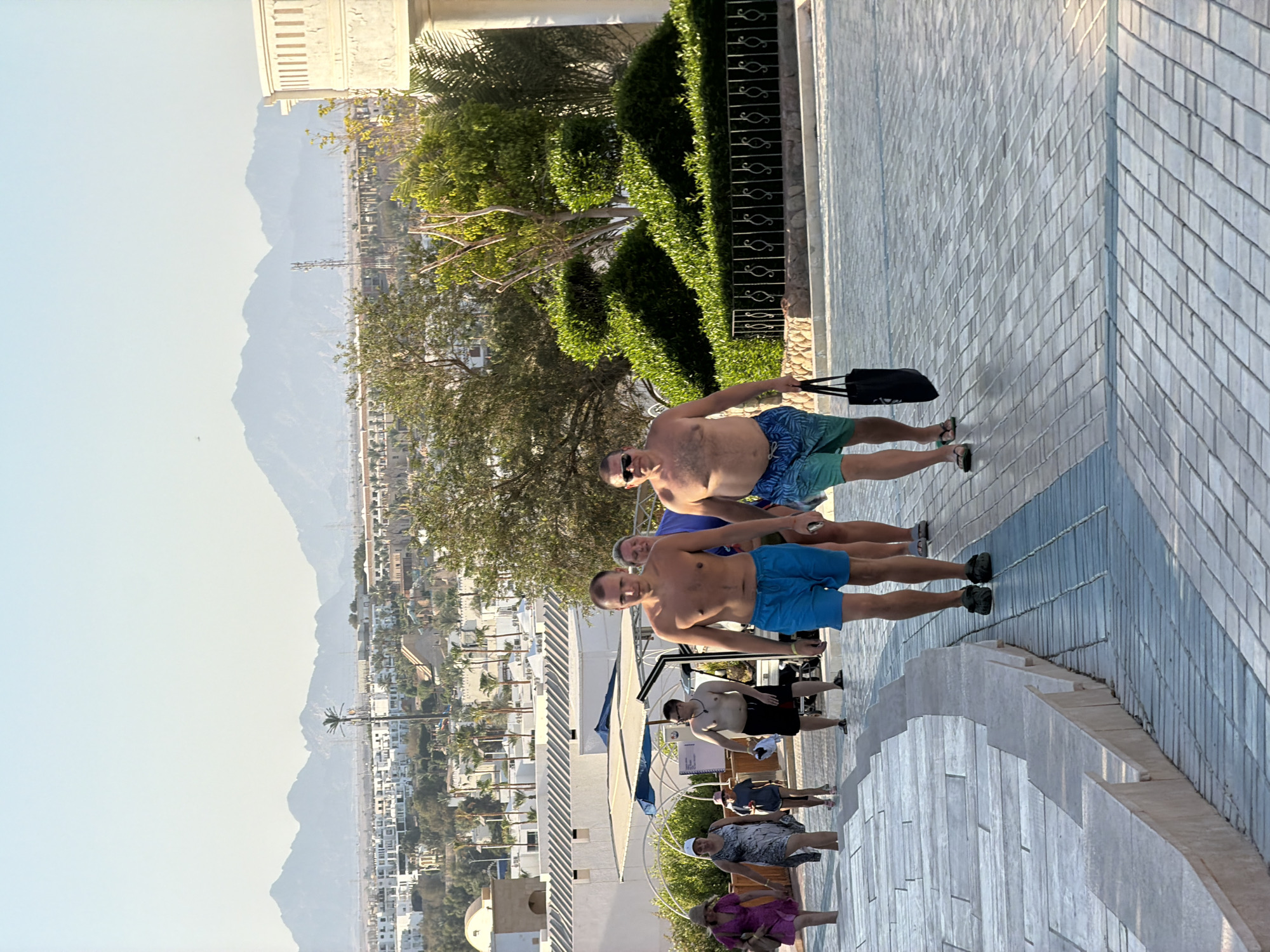 The image depicts a group of people walking on a paved walkway in Sharm El Sheikh, Egypt. In the foreground, two men, likely tourists, are walking towards the viewer. The man on the left wears blue swim trunks and is barefoot. The man on the right wears blue and green patterned swim trunks and sunglasses, and carries a black tote bag. Behind them, other individuals are also walking up a sloped walkway. In the mid-ground, a golf cart is parked near a shaded structure. Beyond the walkway, white buildings with arched windows and palm trees are visible, indicative of a resort area. In the background, hazy mountains form a scenic backdrop. The lighting suggests it is daytime, possibly late afternoon, with the sun casting long shadows.