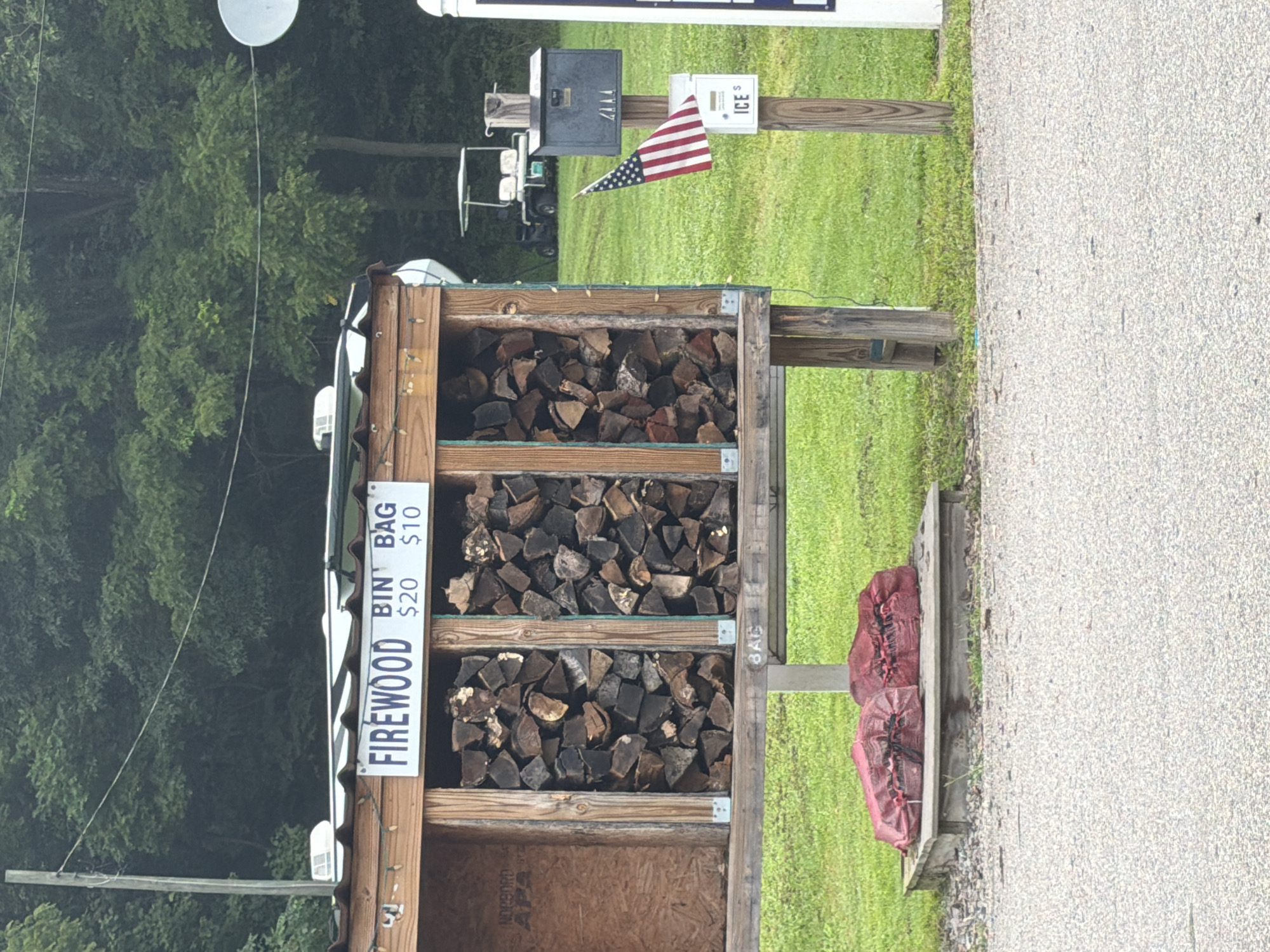 The image depicts a roadside firewood stand in South Beaver Township, United States of America. The stand is made of wood and has a white, corrugated metal roof adorned with string lights. A sign above the firewood reads "FIREWOOD BIN BAG $20 $10." The stand is filled with split logs. To the left of the firewood bin, there are two red bags of what appears to be more firewood. In the background, there is a grassy area with trees. A golf cart, a mailbox with a small American flag attached, and a white sign with the word "ICE" are also visible. The ground in front of the stand is covered with gravel and asphalt.