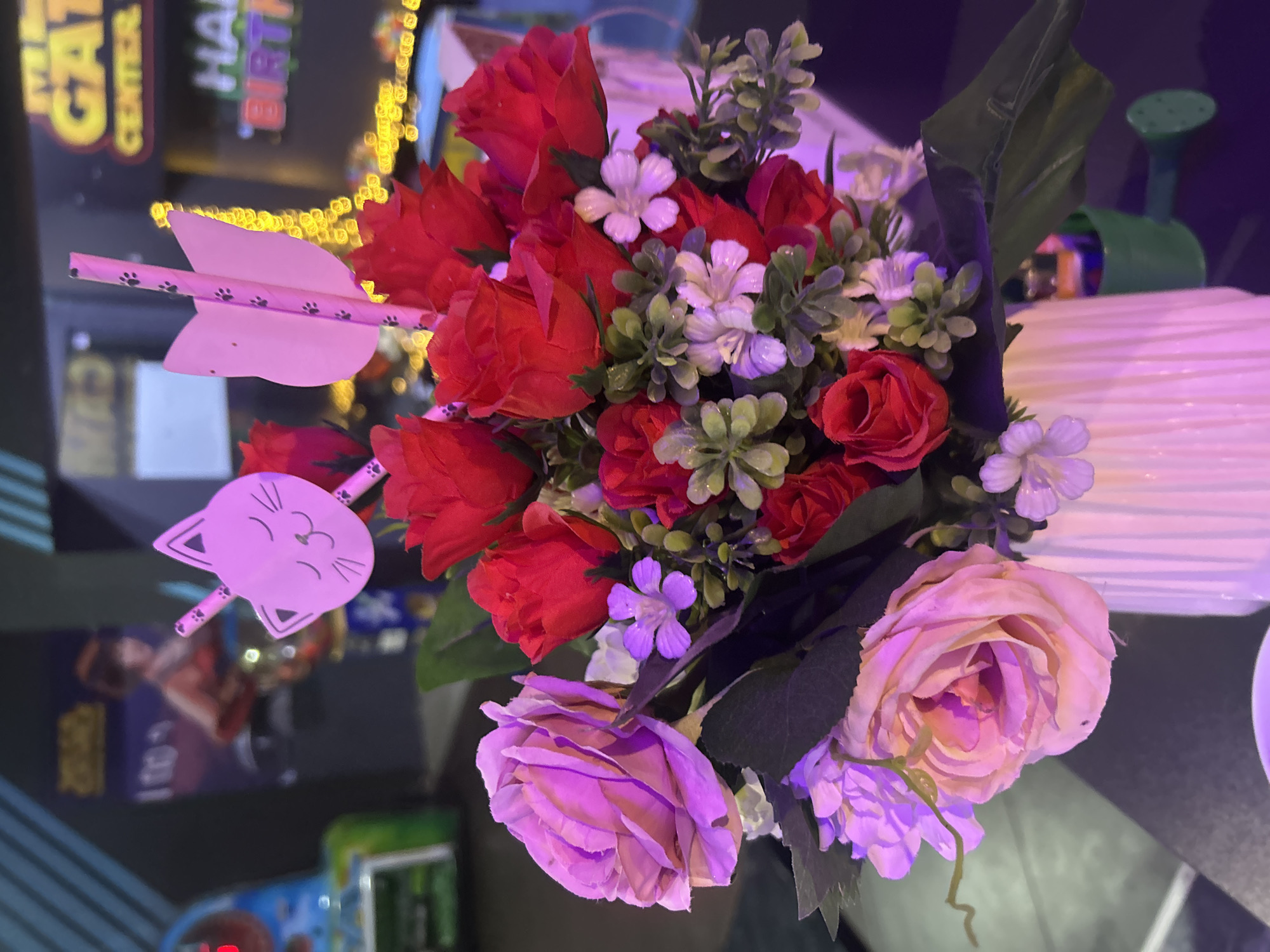 This image shows a close-up view of a floral arrangement in a white fluted vase. The bouquet is composed of several red roses, two large light pink roses, and smaller white and purple flowers, interspersed with green artificial foliage. Two pink decorative straws, one shaped like a cat's face and the other with a bow-like shape, are inserted among the flowers. The background is blurred but suggests an indoor entertainment venue, possibly an arcade or amusement center, with colorful lights and signage visible. One sign appears to say "GAT CENTER" and another "HAPPY BIRTHDAY". The overall lighting is dim with a purple hue, likely from ambient lighting or a filter. The scene is likely located in Burgas, Bulgaria, given the context of the request.