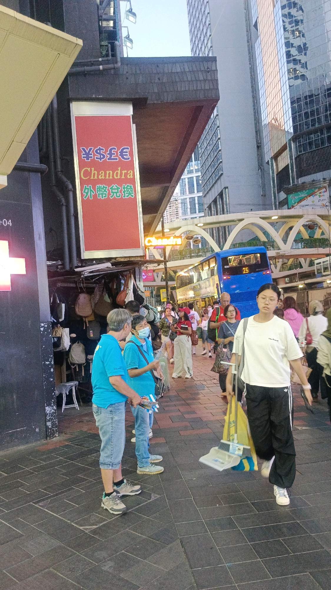 The image depicts a street scene in a busy urban environment, likely Hong Kong, China, given the presence of Chinese characters and a double-decker bus. In the foreground, two individuals in blue shirts are standing on a paved sidewalk. One is holding a plastic bottle, and the other is holding some papers. To their right, a person in a white t-shirt and black track pants is sweeping the ground with a broom and dustpan. Behind them, a blue double-decker bus with the number "25" displayed on the front is passing by. The background features modern high-rise buildings with glass facades, indicating a commercial or financial district. A red sign with foreign currency symbols (¥$£) and Chinese text is visible, suggesting a currency exchange service. Several other people are walking in the background, and bags are displayed for sale at a stall. The lighting suggests it is late afternoon or early evening.
