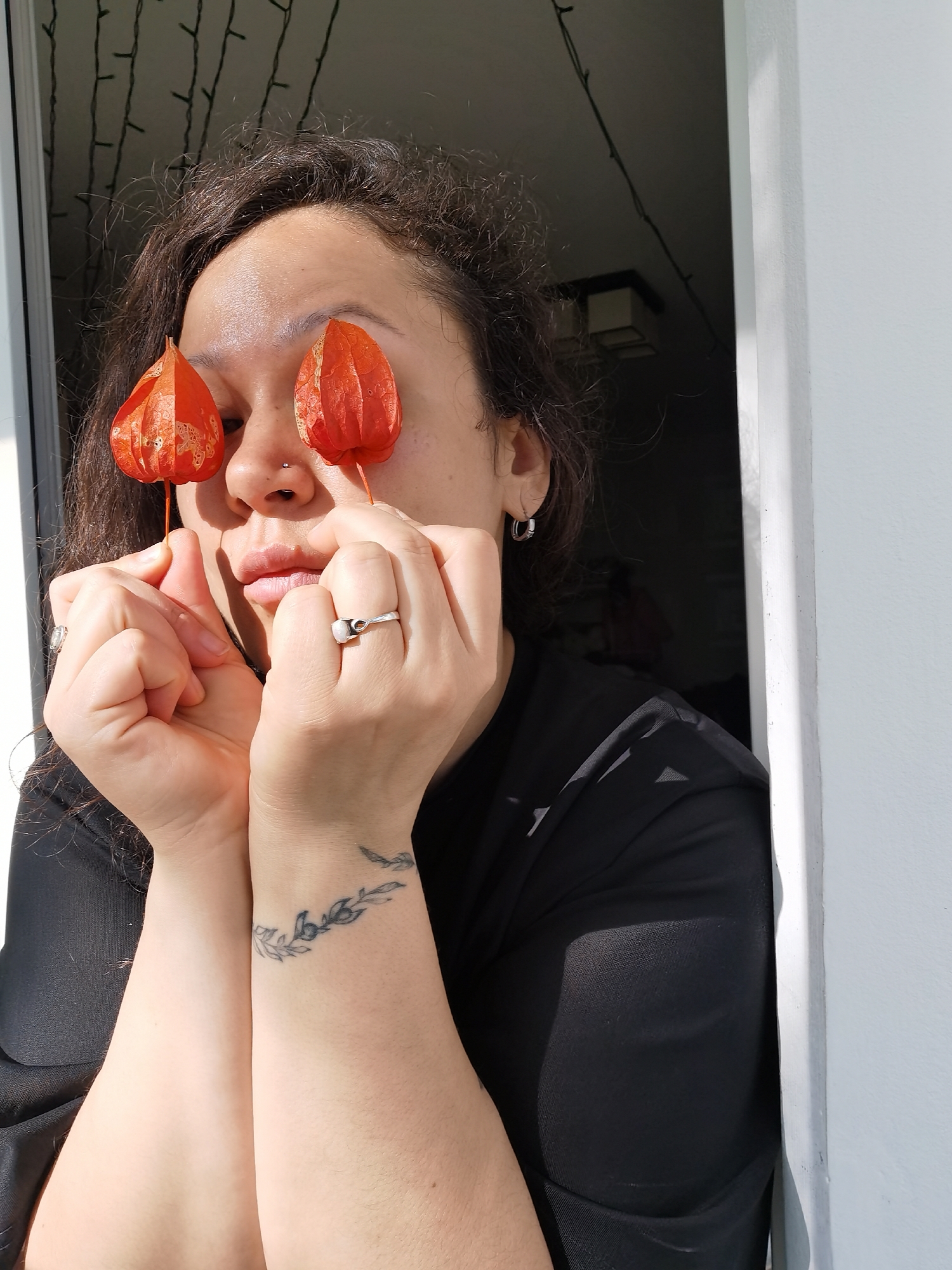 A woman is positioned in front of an open window, holding two orange lantern-shaped flowers over her eyes, effectively obscuring her vision. The woman is fair-skinned with dark, curly hair. She has a nose piercing and is wearing a ring on her left hand and hoop earrings. Her arms are raised and bent, with her hands close to her face. A tattoo is visible on her left wrist. She is wearing a black shirt. The background shows a dark interior and the white frame of the open window. String lights can be seen in the background. The location is Chaikovsky, Russia.