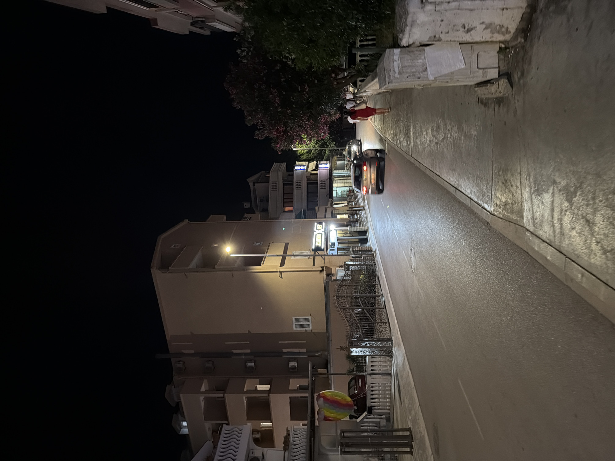 This image depicts a street scene at night in the city of Budva, Montenegro. A multistory building with balconies and air conditioning units lines the left side of the street. In front of this building, a decorative rainbow-colored balloon is visible. A black metal gate with ornate detailing separates the property from the sidewalk. Across the street, another multistory building features a lit sign that appears to read "GELATO CITY."
The road in the foreground is paved and wet, reflecting the ambient light from streetlights and building facades. A silver car is seen driving away from the viewer on the right side of the road, its taillights illuminated. On the sidewalk to the right, a woman wearing a red dress is walking away from the viewer. Behind her, a dark-colored car is parked. Further down the sidewalk, several other individuals are walking in the same direction as the woman in the red dress. Trees with pink blossoms are visible in the background, bordering the sidewalk. The sky is dark, indicating it is nighttime. The scene suggests a commercial or residential street with pedestrian activity.